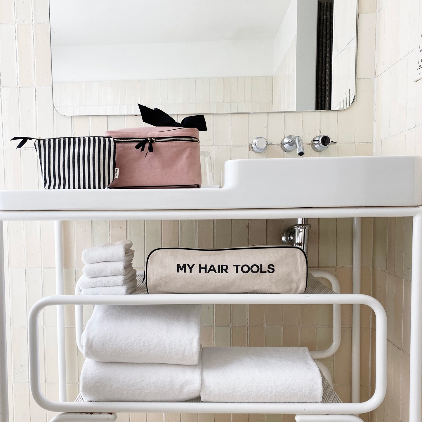 Bag-all Roomy Hair Tools Travel Case in cream displayed on modern bathroom shelf with white towels, striped cosmetic bag, featuring 'My Hair Tools' text and heat-resistant lining