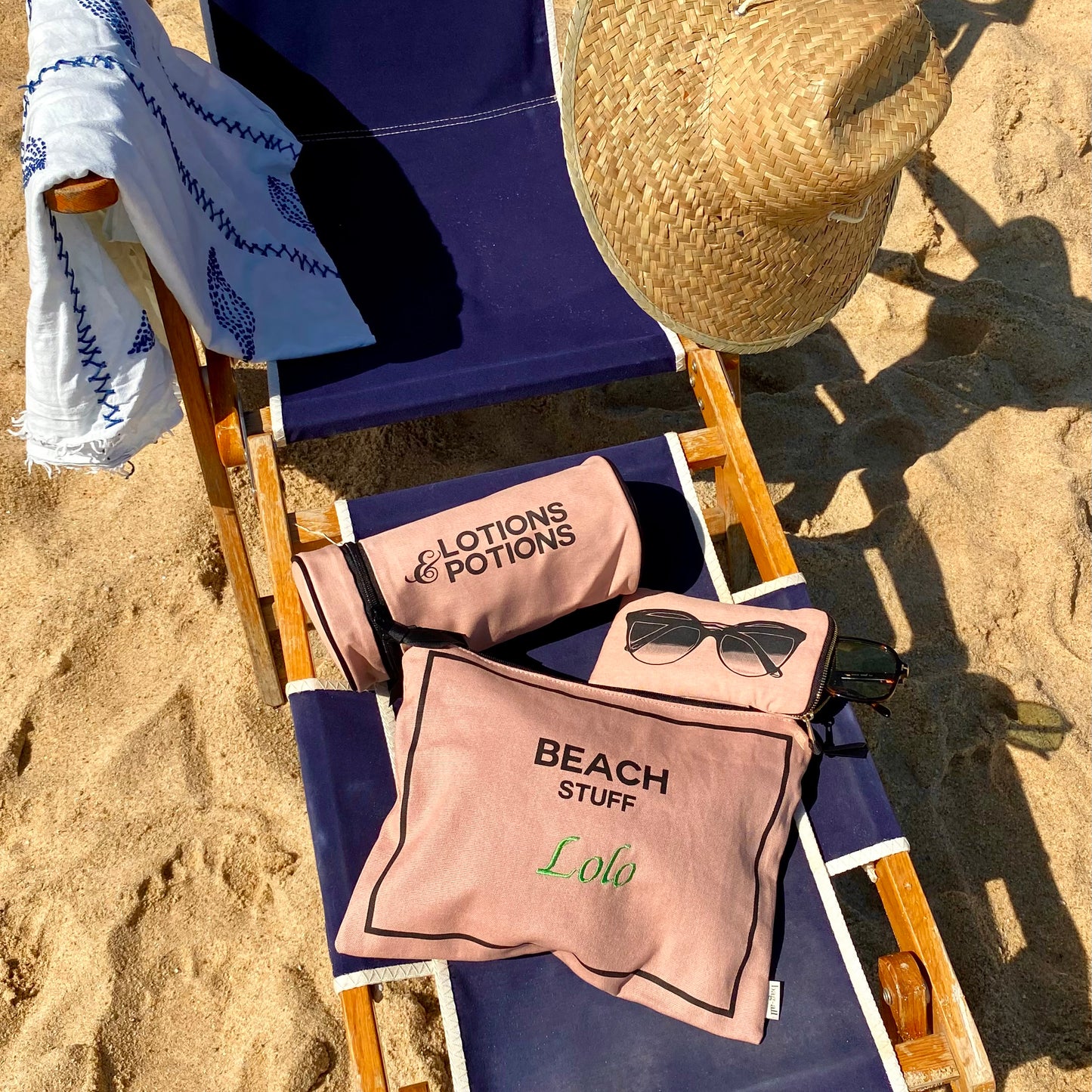 Bag-all Stylish Beach Essential Set in pink/blush featuring three monogrammable cotton pouches for beach items, sunglasses, and lotions, arranged on blue beach chair with straw hat in sand