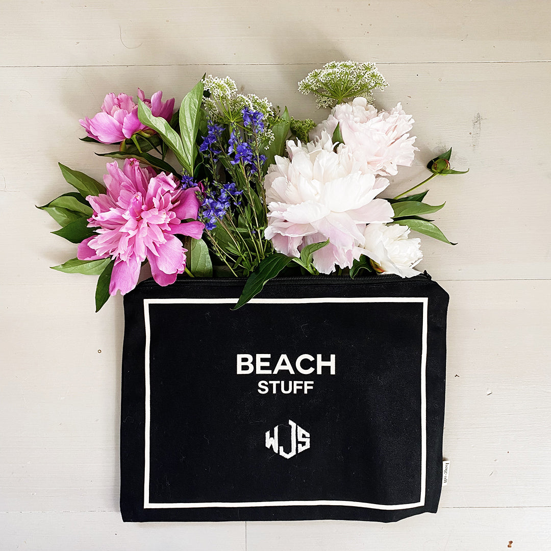 Bag-all Monogram Beach Pouch in black cotton with white border trim, displaying "BEACH STUFF" text and monogram, styled with pink and white peonies and blue wildflowers