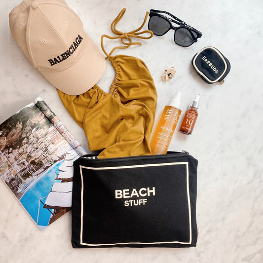 Bag-all Beach Pouch in black cotton with waterproof lining, featuring "BEACH STUFF" text and zipper closure, displayed with summer essentials including sunscreen, swimsuit, and beach accessories