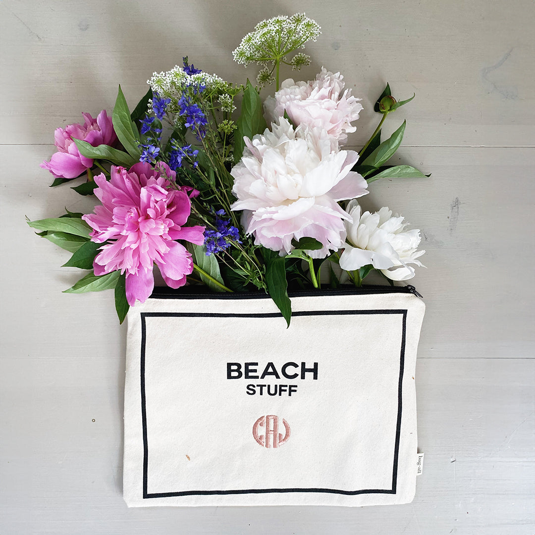 Bag-all Monogram Beach Pouch in cream canvas with 'Beach Stuff' text, filled with pink and white peonies, blue delphinium, and white Queen Anne's lace flowers, displaying elegant storage solution