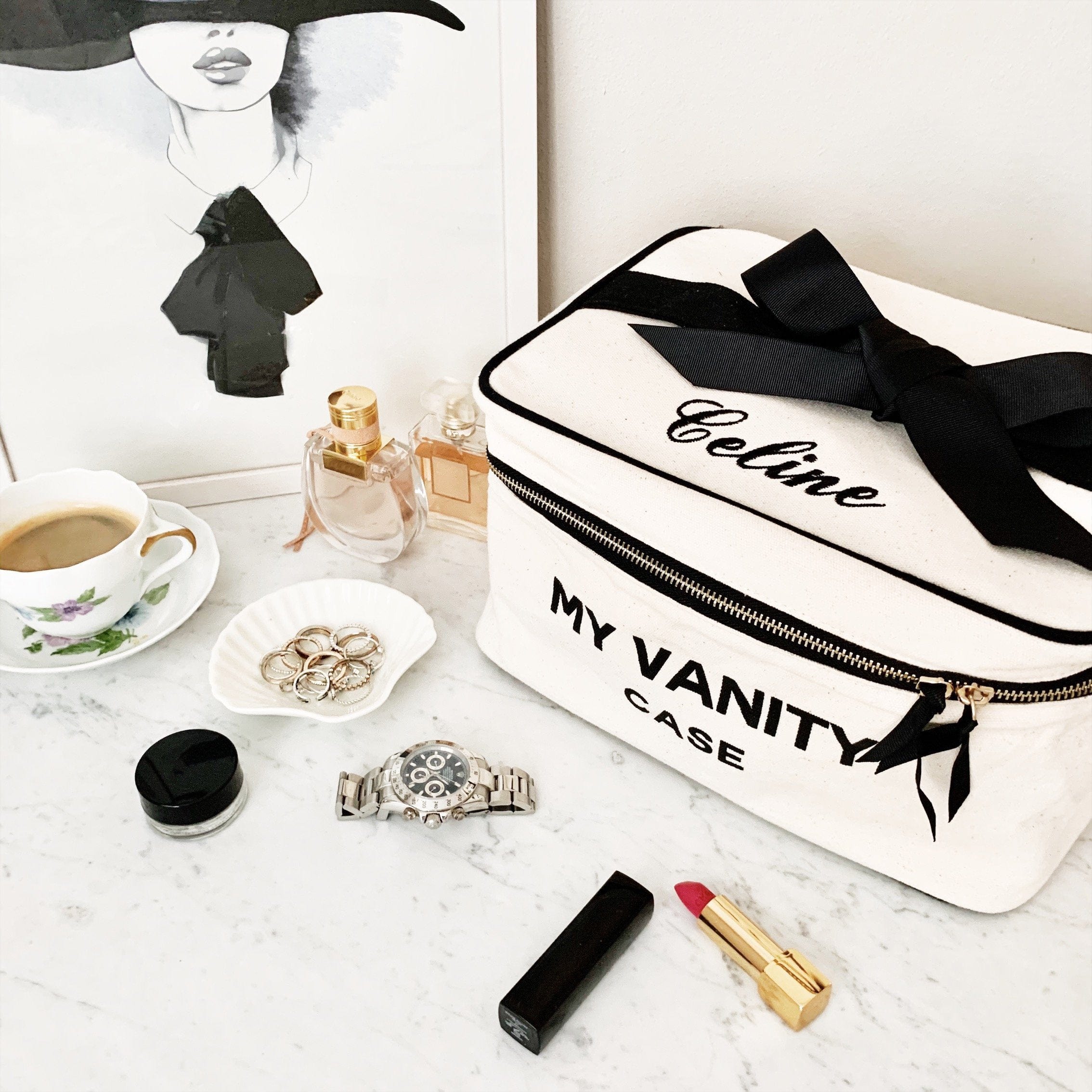 Cream Bag-all vanity case with black ribbon and zipper on marble table with lipstick, watch, rings, perfume, and coffee cup