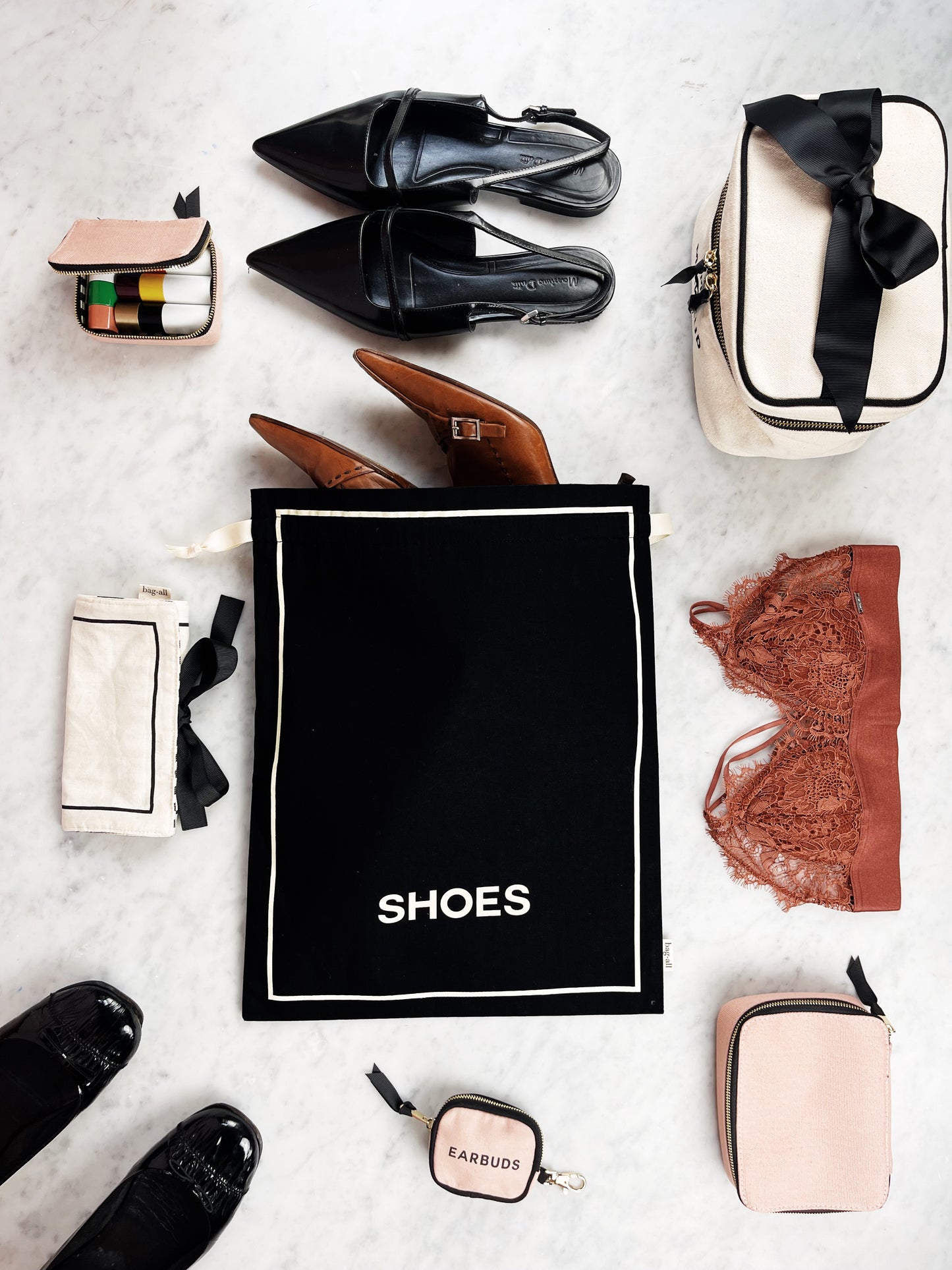 Bag-all Couture Shoe Travel Bag in black with white border, displayed with luxury accessories including leather shoes, lingerie, and travel essentials on marble surface