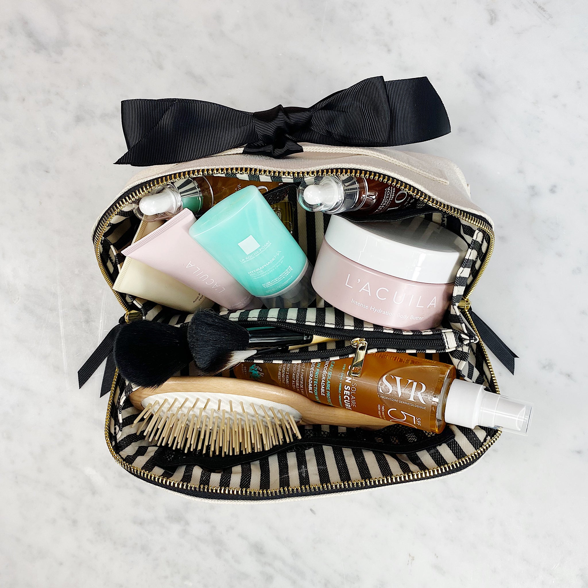 Bag-all Beauty Organizer Case with striped interior, displaying skincare products, hairbrush, and black bow detail. Perfect for storing cosmetics and travel essentials.