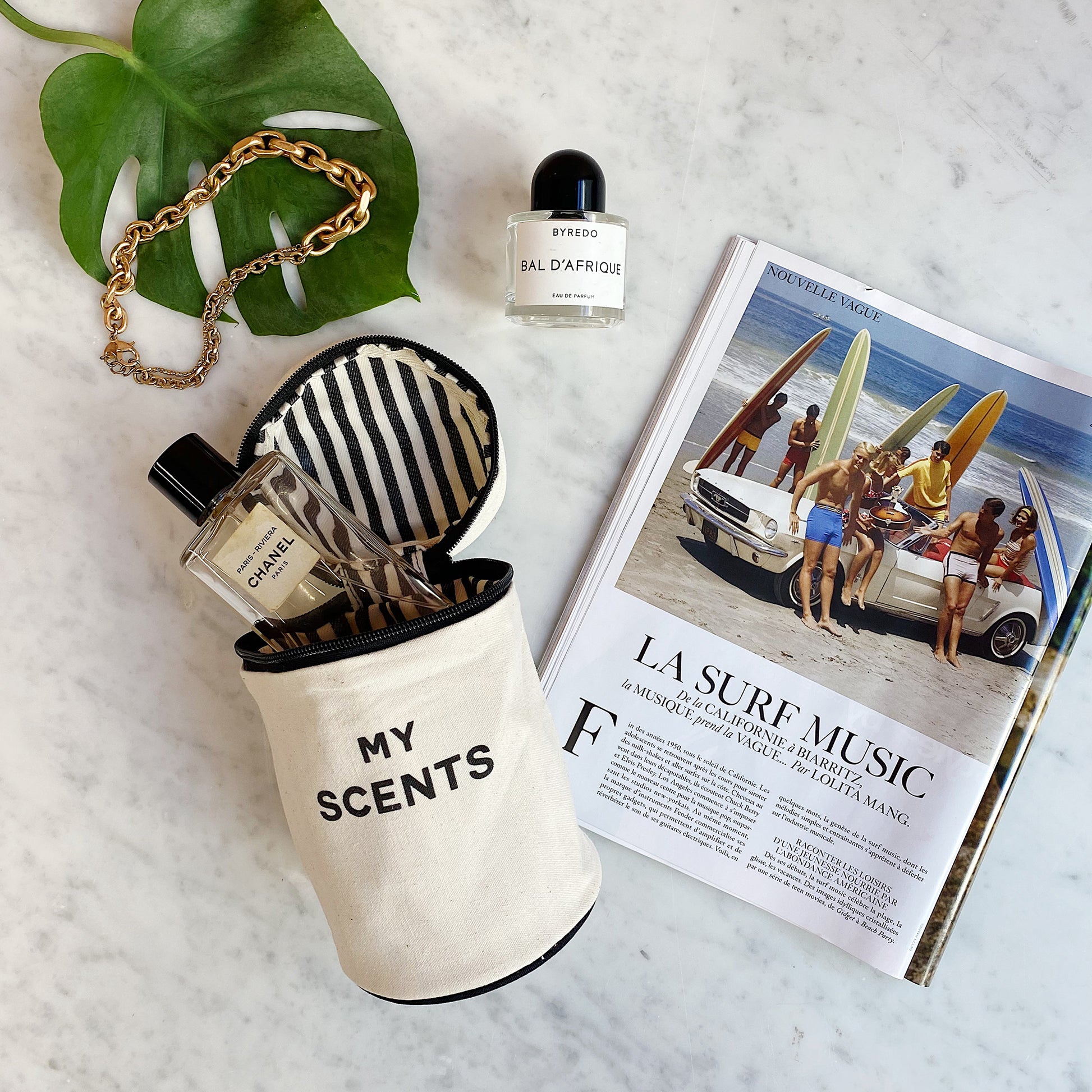 Bag-all Round My Scents Case in cream with black striped interior, displaying perfume bottles, next to a fashion magazine and decorative leaf