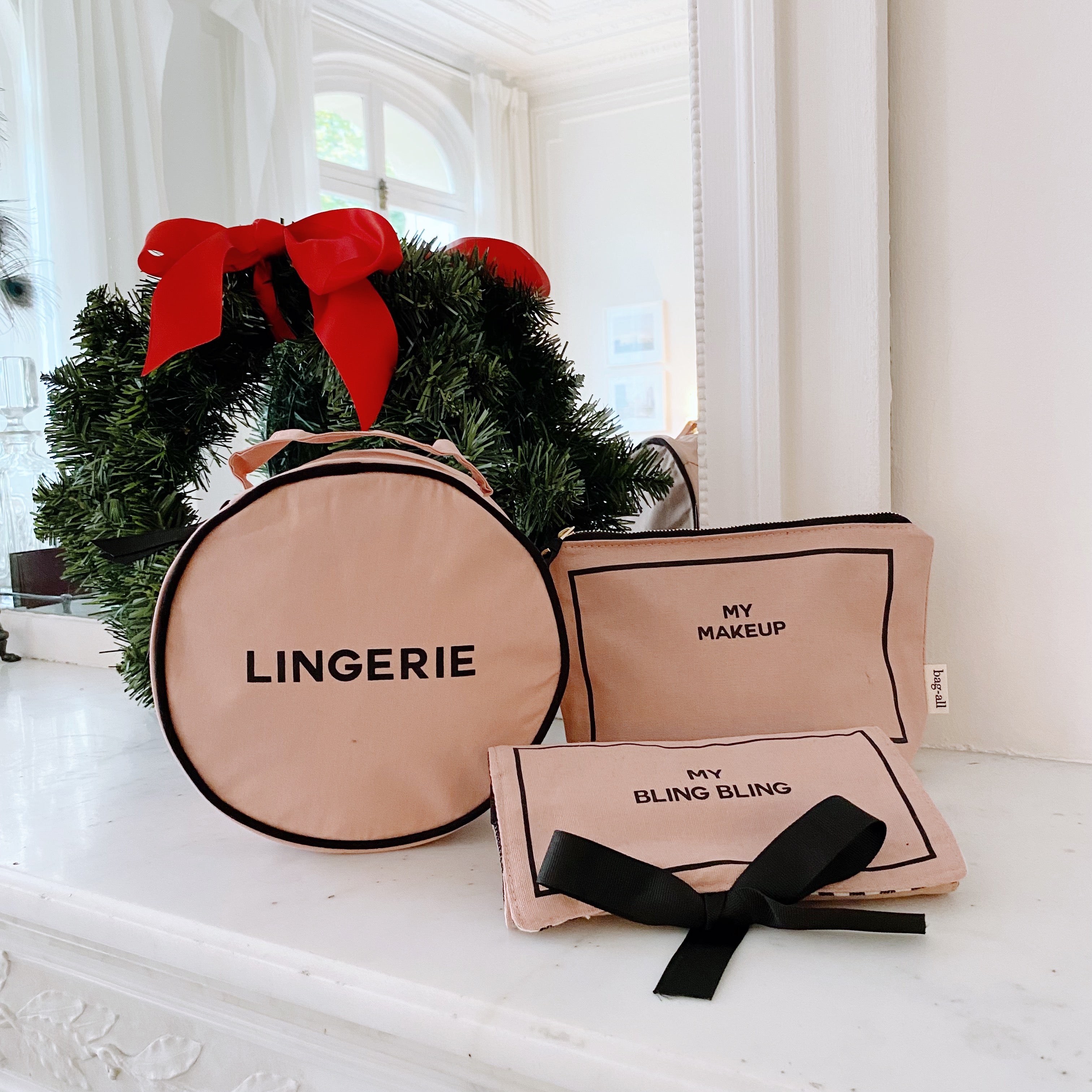 Pink and blush Bag-all 3-pack set with round lingerie case, makeup case, and jewelry pouch with black bow near Christmas wreath