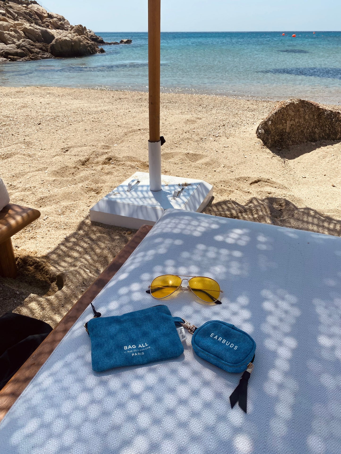 Bag-all Denim Earbuds Case with Gold Clasp shown on beach lounger alongside yellow sunglasses, showcasing its compact size and stylish blue design perfect for summer beach accessories and travel organization