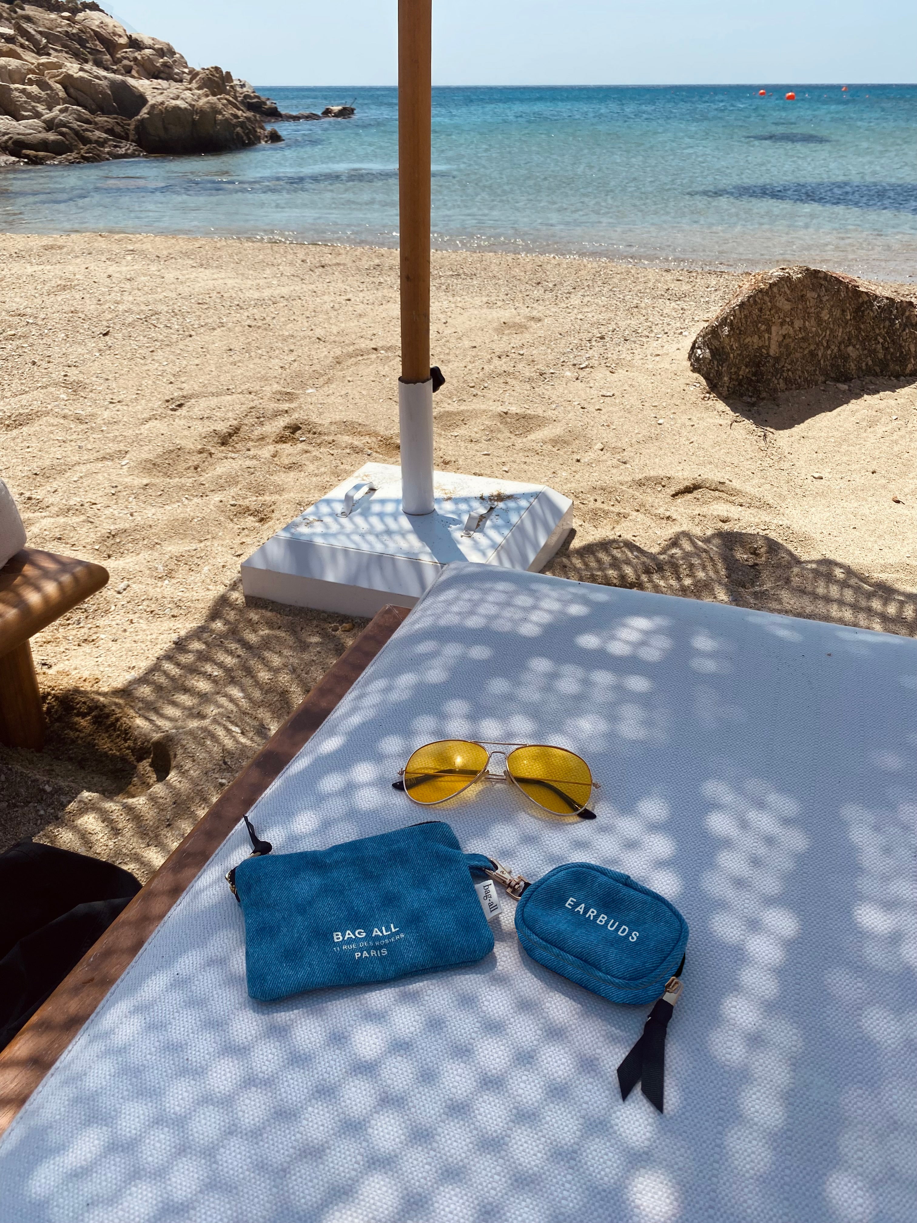 Bag-all Denim Earbuds Case with Gold Clasp shown on beach lounger alongside yellow sunglasses, showcasing its compact size and stylish blue design perfect for summer beach accessories and travel organization