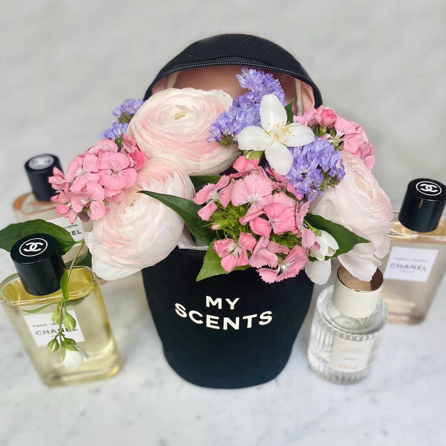 Bag-all Round My Scents Case in black featuring pink ranunculus, purple flowers, and white blooms, displayed with luxury perfume bottles on marble surface, showcasing elegant fragrance storage