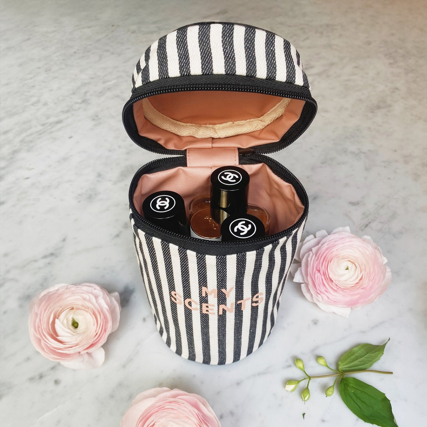 Bag-all Round My Scents Case in black and white stripes, cylindrical perfume organizer with pink interior, shown open displaying perfume bottles on marble surface with pink ranunculus flowers