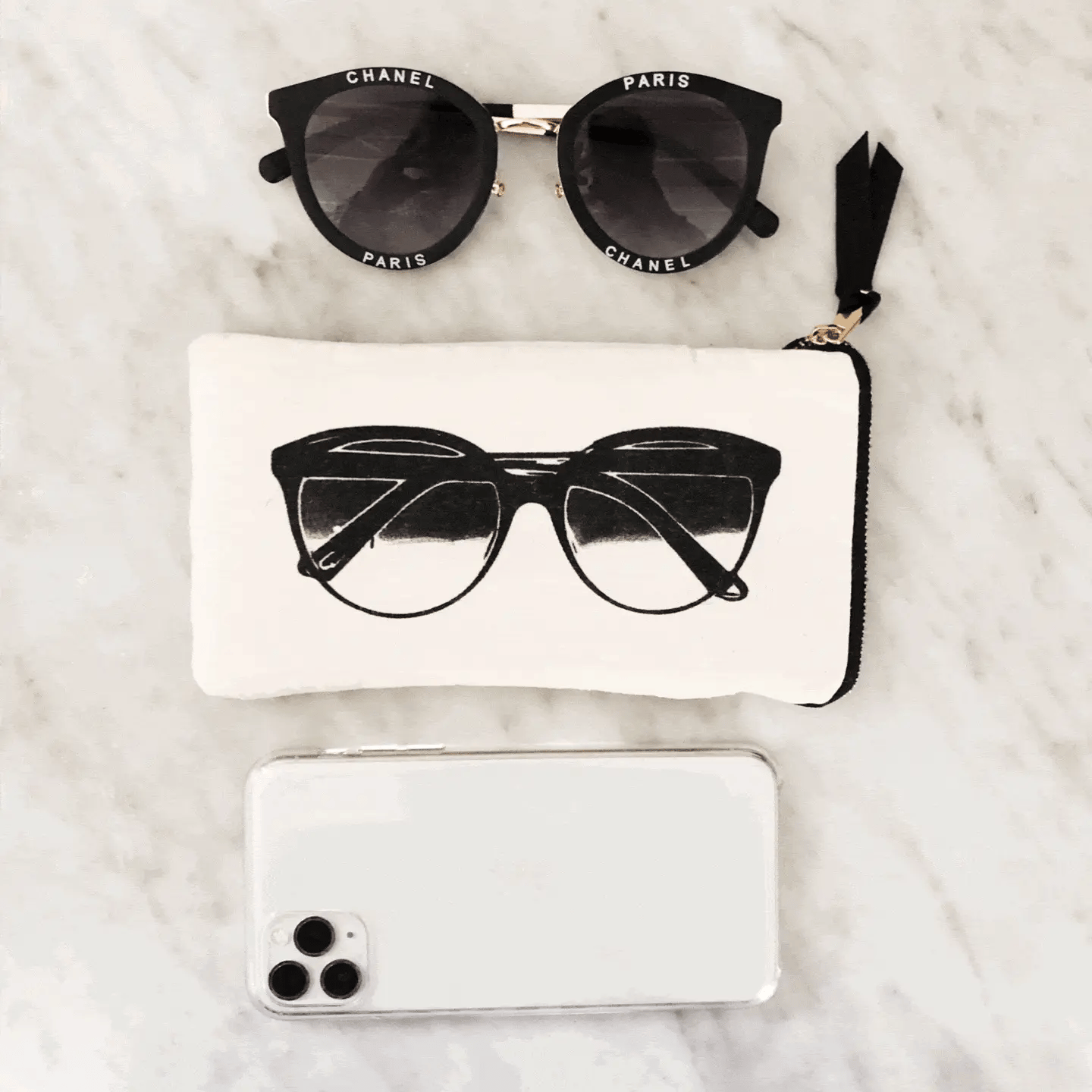 Bag-all Glasses Case with Outside Pocket in Cream featuring black sunglasses print, shown with designer sunglasses and phone on marble surface, stylish protective storage solution