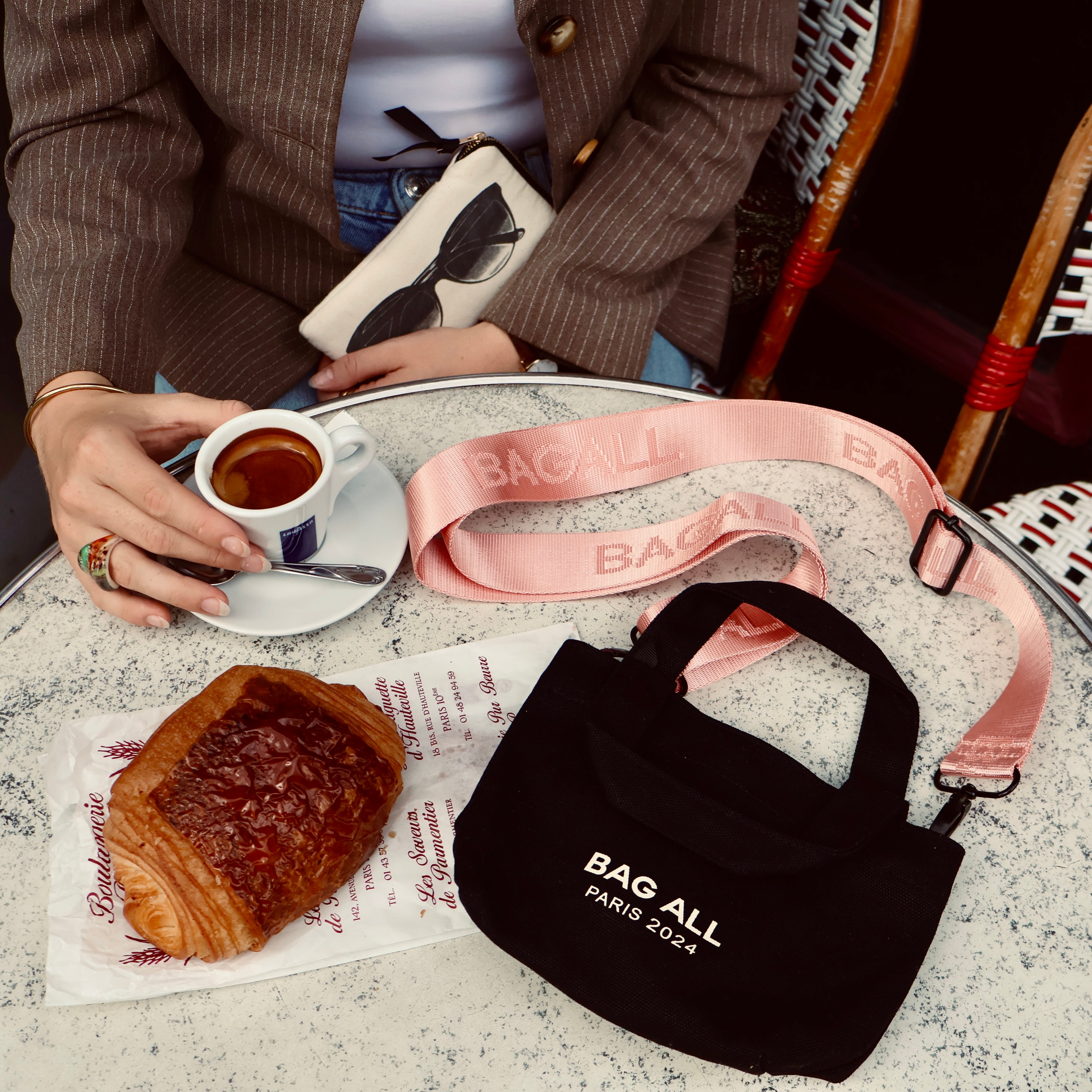 Bag-all Mini Tote Bag in black with adjustable pink strap displayed at cafe setting with coffee and pastry, showcasing versatile everyday use and urban lifestyle appeal