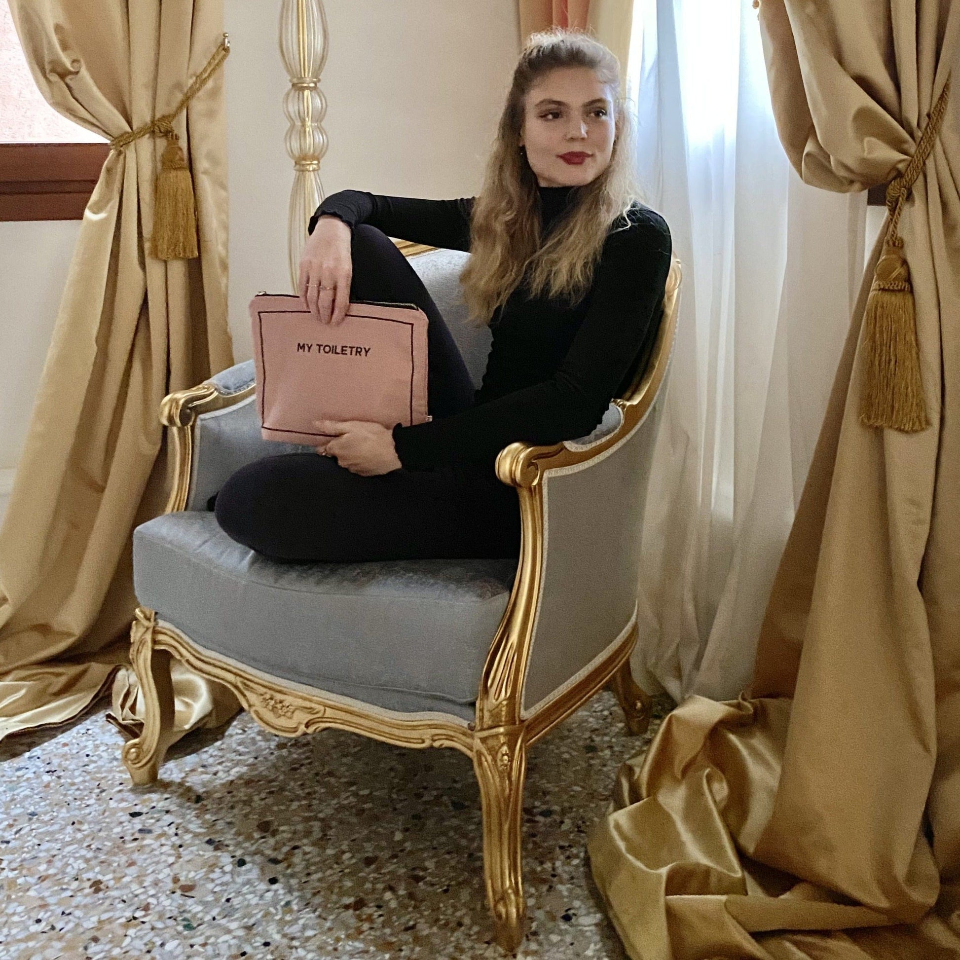 Bag-all Weekend Essential Packing Set shown in elegant setting, featuring pink toiletry pouch on vintage gold-trimmed chair with beige curtains