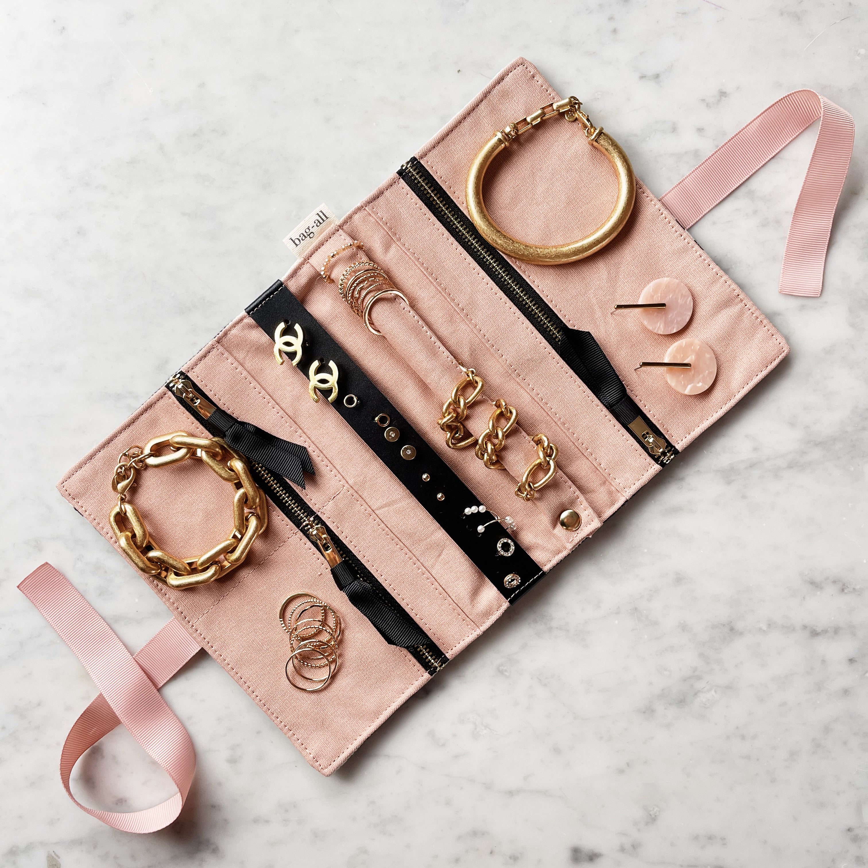 Bag-all Couture Jewelry Roll Medium Striped in pink with black zippers, featuring multiple compartments displaying gold jewelry and accessories on marble background
