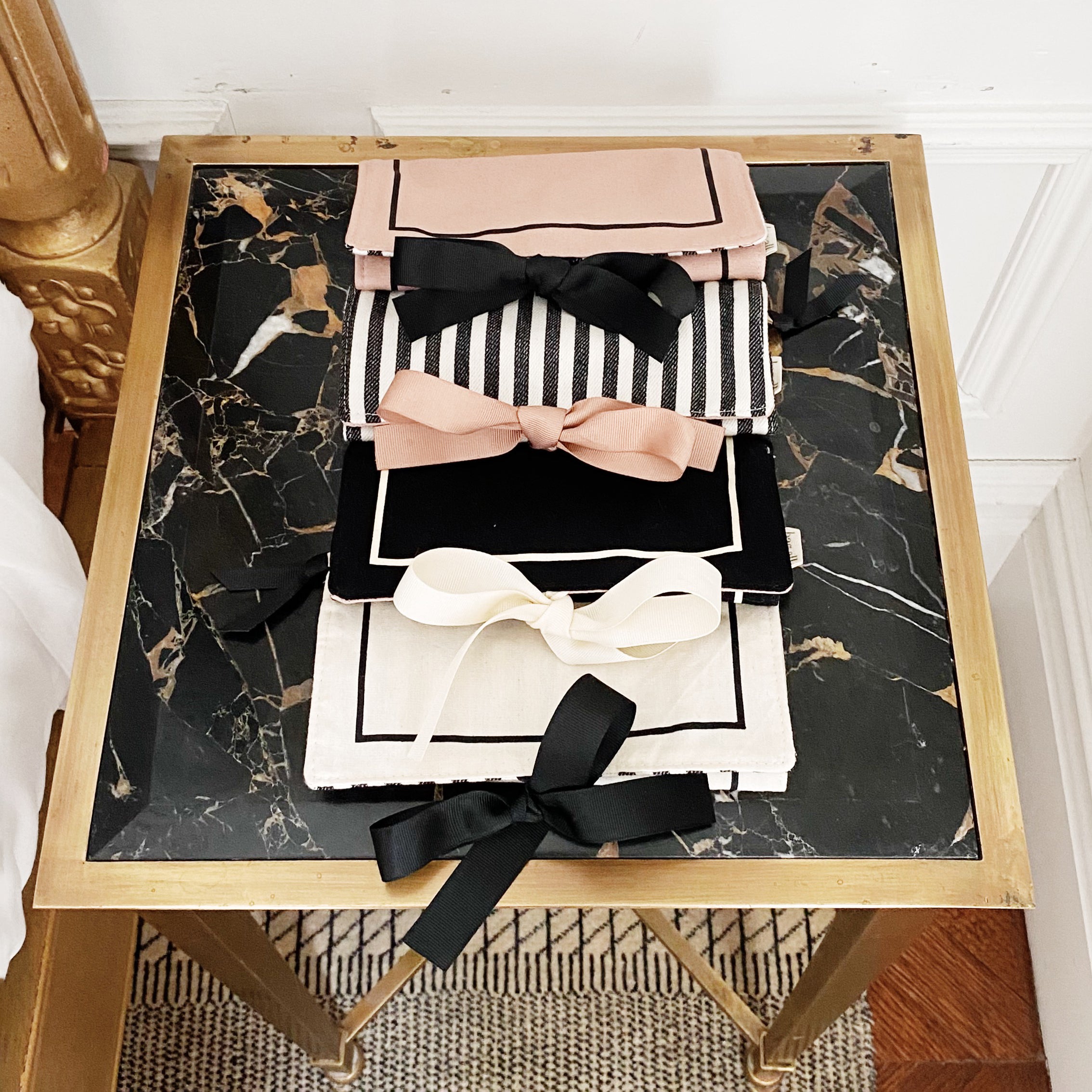 Bag-all Couture Jewelry Roll Medium Black, featuring elegant ribbon-tied travel organizers in black, pink, and white stacked on marble-top table, showcasing stylish storage solutions