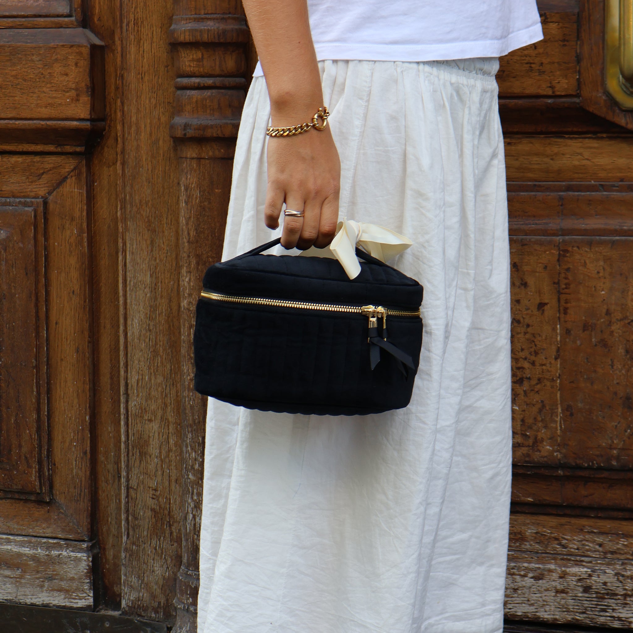 Woman holding a black quilted eco velvet makeup case with gold zipper from Bag-all against wooden door background
