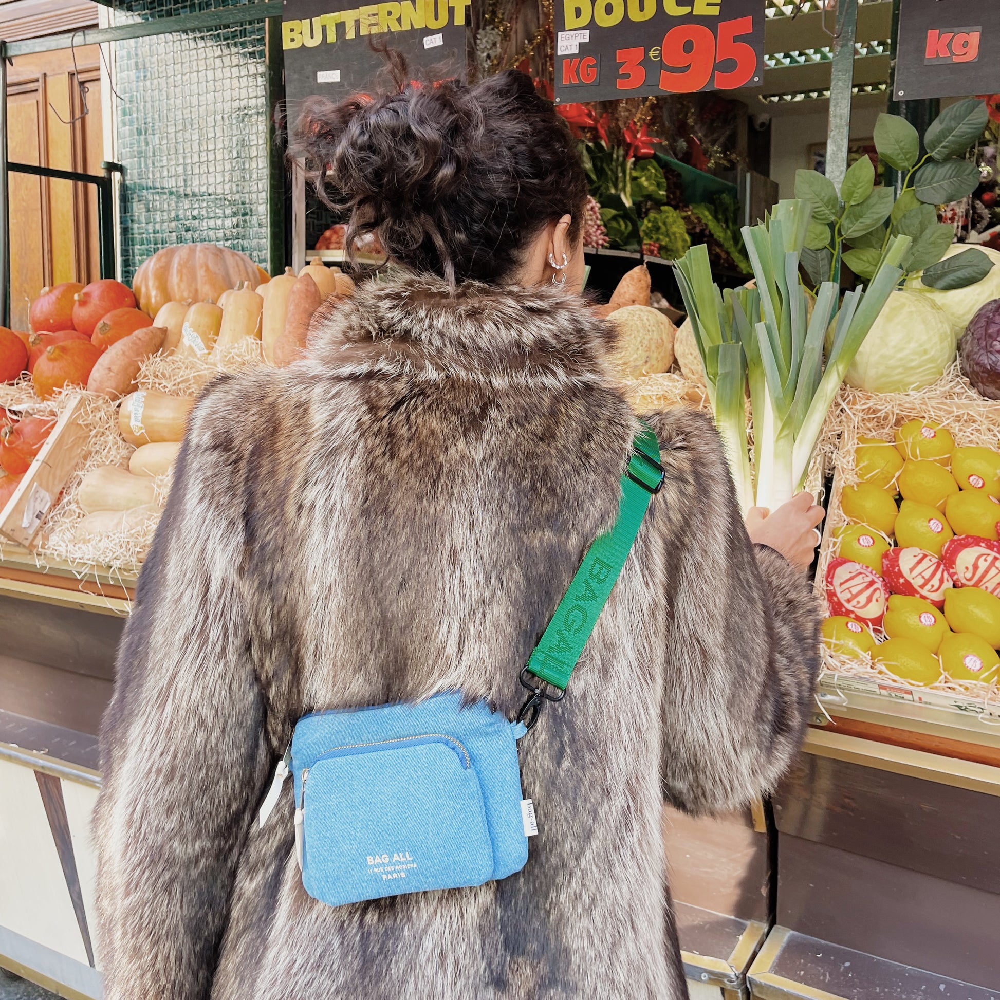 Bag-all Padded Trinket Pouch Double Pocket in denim blue with green strap worn crossbody, shown at produce market. Versatile organizing pouch features two compartments and padded protection.