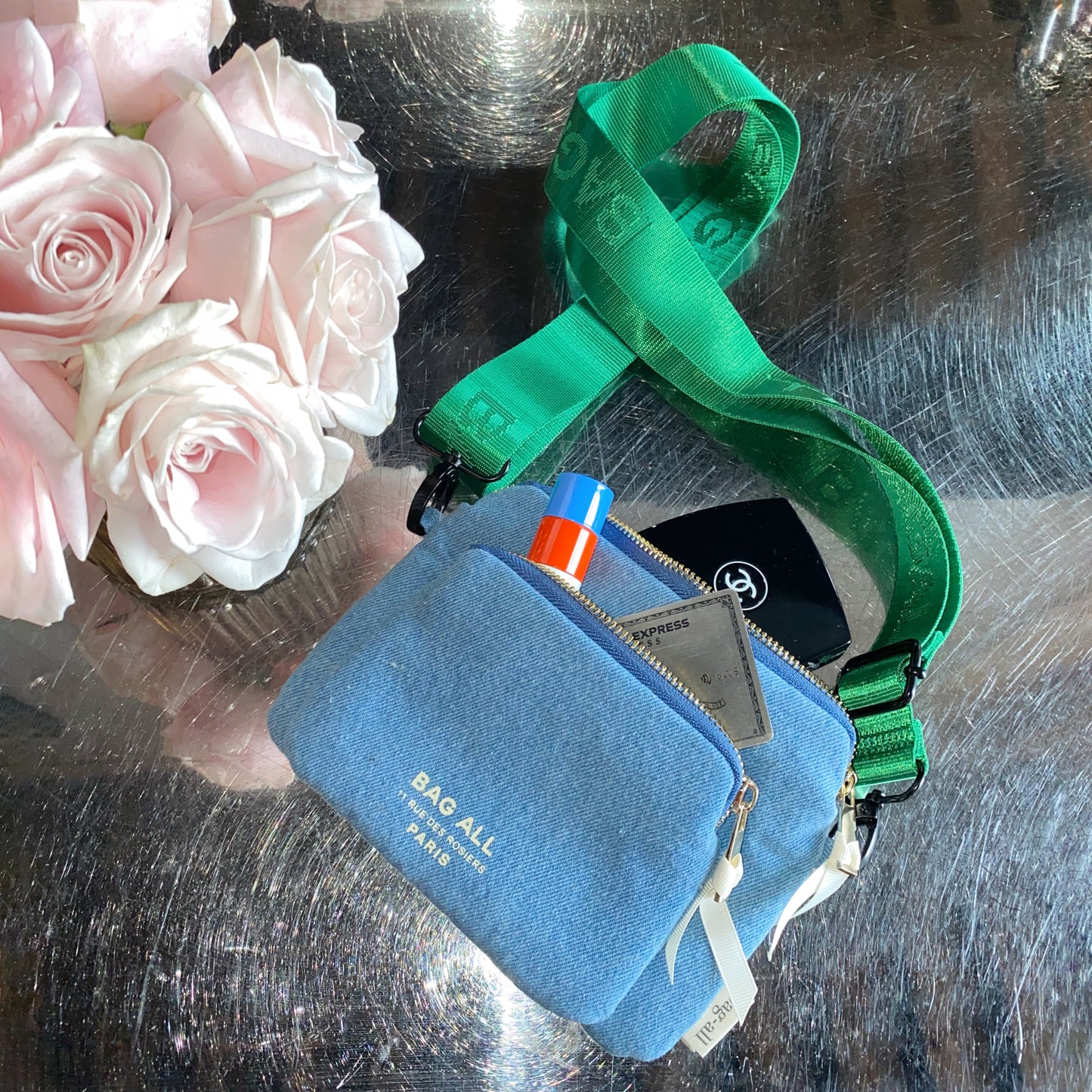 Bag-all Padded Trinket Pouch Double Pocket Denim with green adjustable strap, displayed next to pink roses. Features dual zipper compartments for organizing small essentials and valuables.