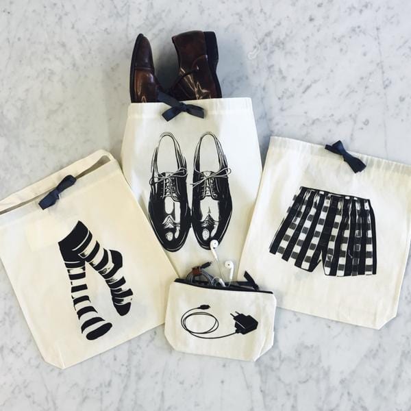Bag-all stylish organizing bags featuring black and white illustrations of dress shoes, striped socks, plaid shorts, and tech accessories on natural cotton pouches with bow details