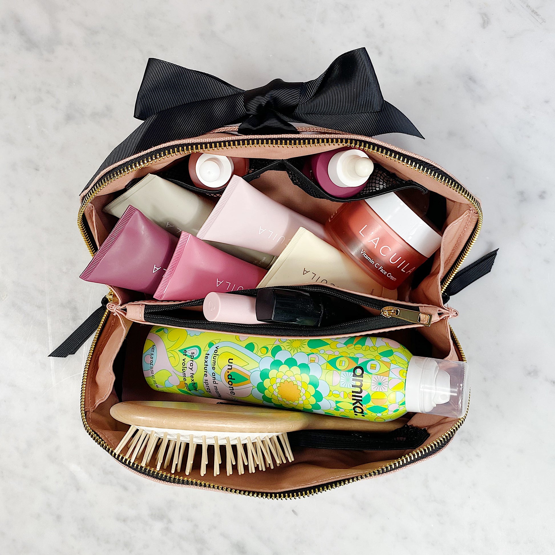 Bag-all stylish beauty organizer case opened to display neatly arranged cosmetics, hair products, and brush with elegant black bow detail and gold zipper against marble surface
