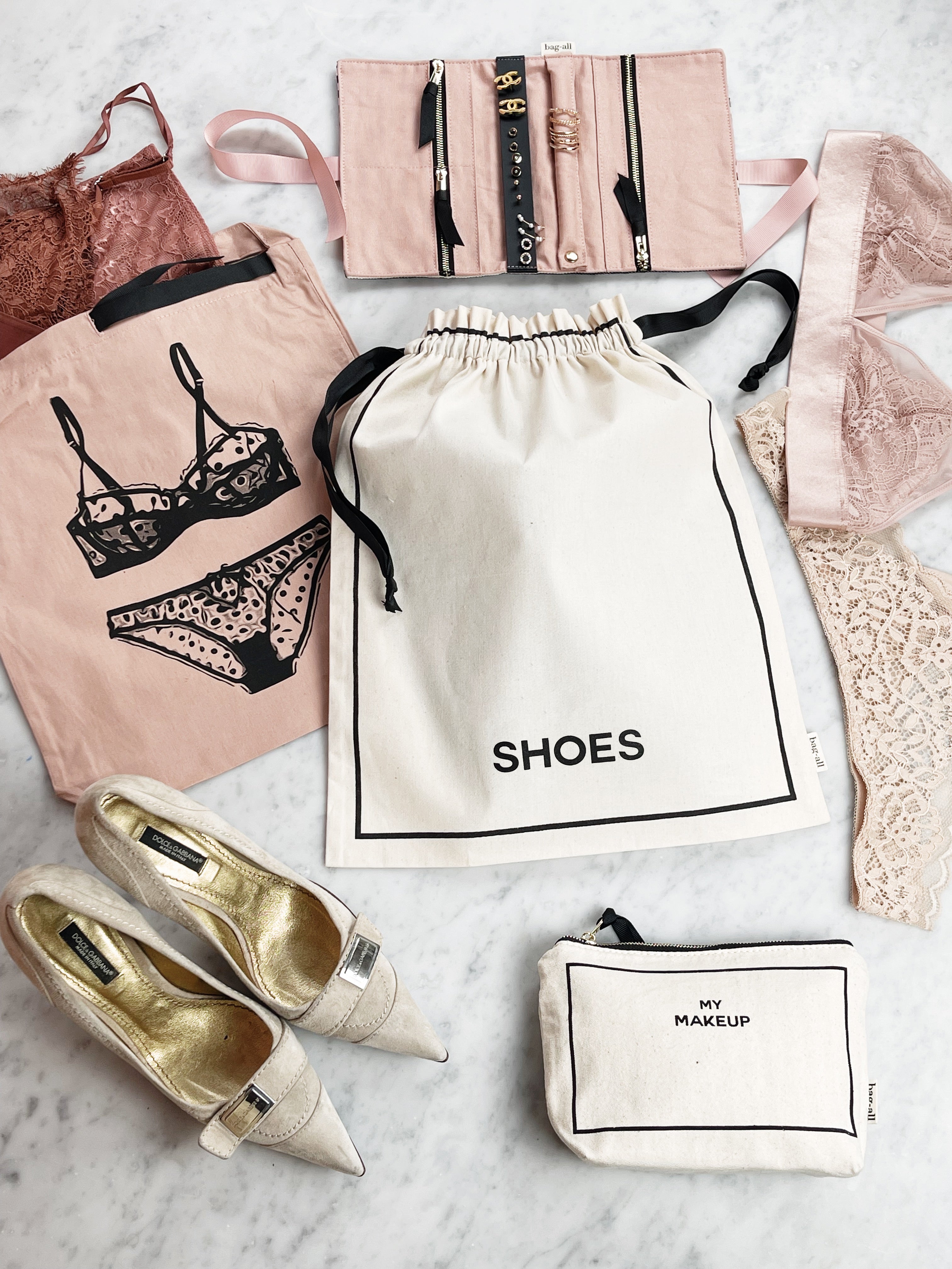 Bag-all Couture Shoe Travel Bag in cream with small print, shown with coordinating travel organizers, gold shoes, and lingerie bags on marble surface, featuring drawstring closure and elegant black text labeling