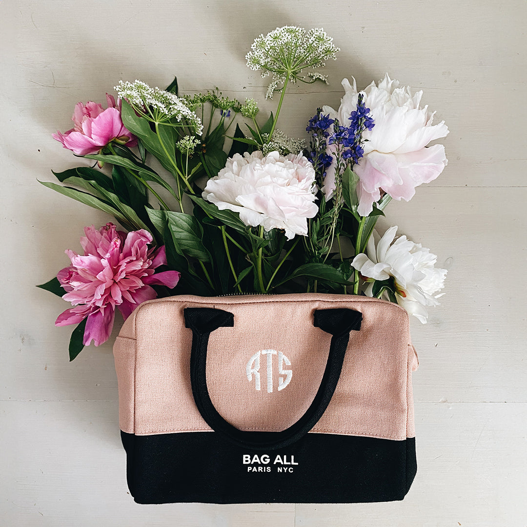 Bag-all Stylish Monogram Lunch Box in pink and black canvas filled with fresh pink peonies, white carnations, delicate Queen Anne's lace, and purple delphiniums against a light background