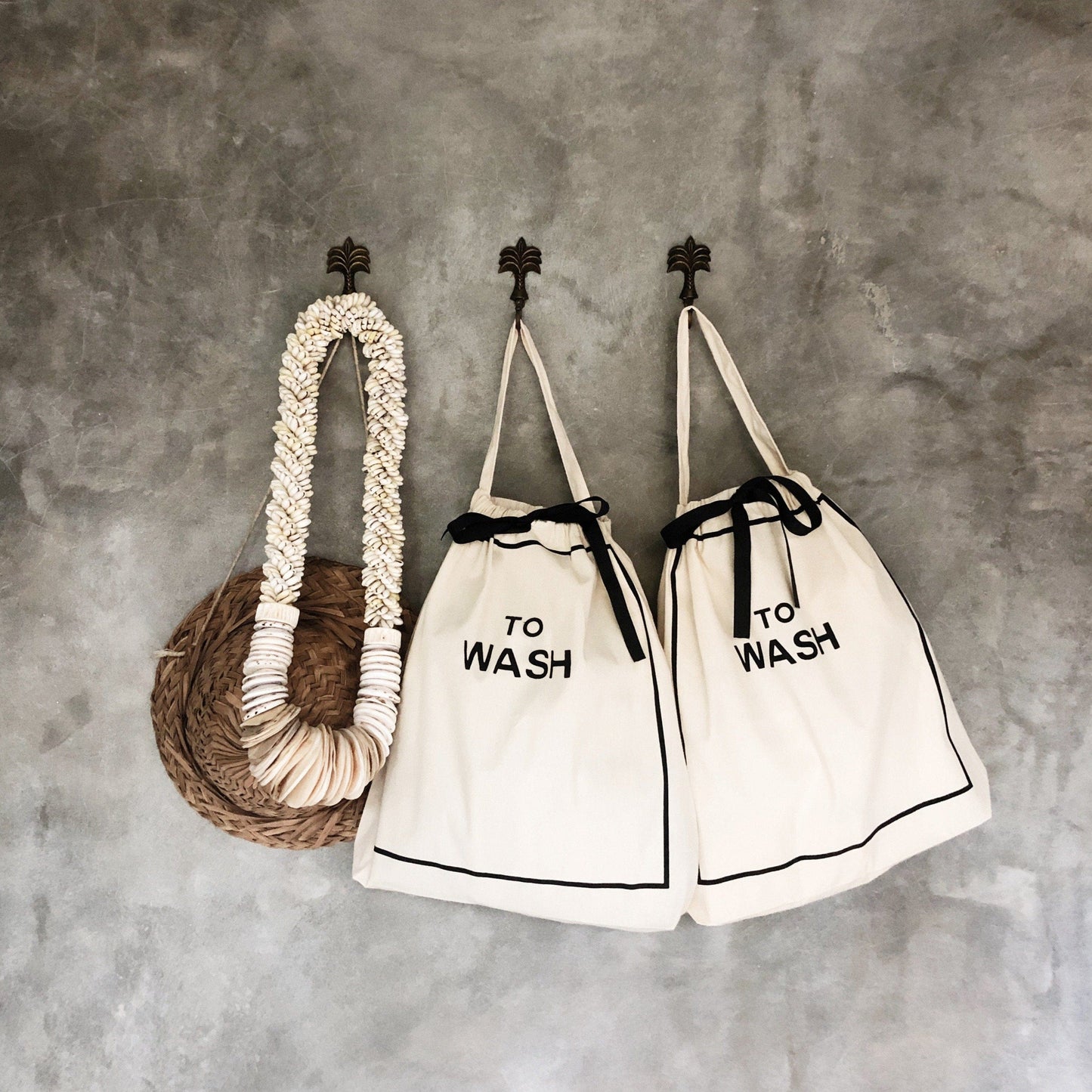 Bag-all To Wash Laundry Bag in cream with black text, hanging on wall hooks alongside matching bag and decorative rope, featuring drawstring closure and minimalist design
