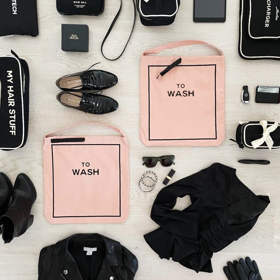 Bag-all To Wash Laundry Bag in pink/blush with black text, displayed among black clothing items, shoes, and accessories on a light background. Stylish cotton organizer for laundry storage and travel.