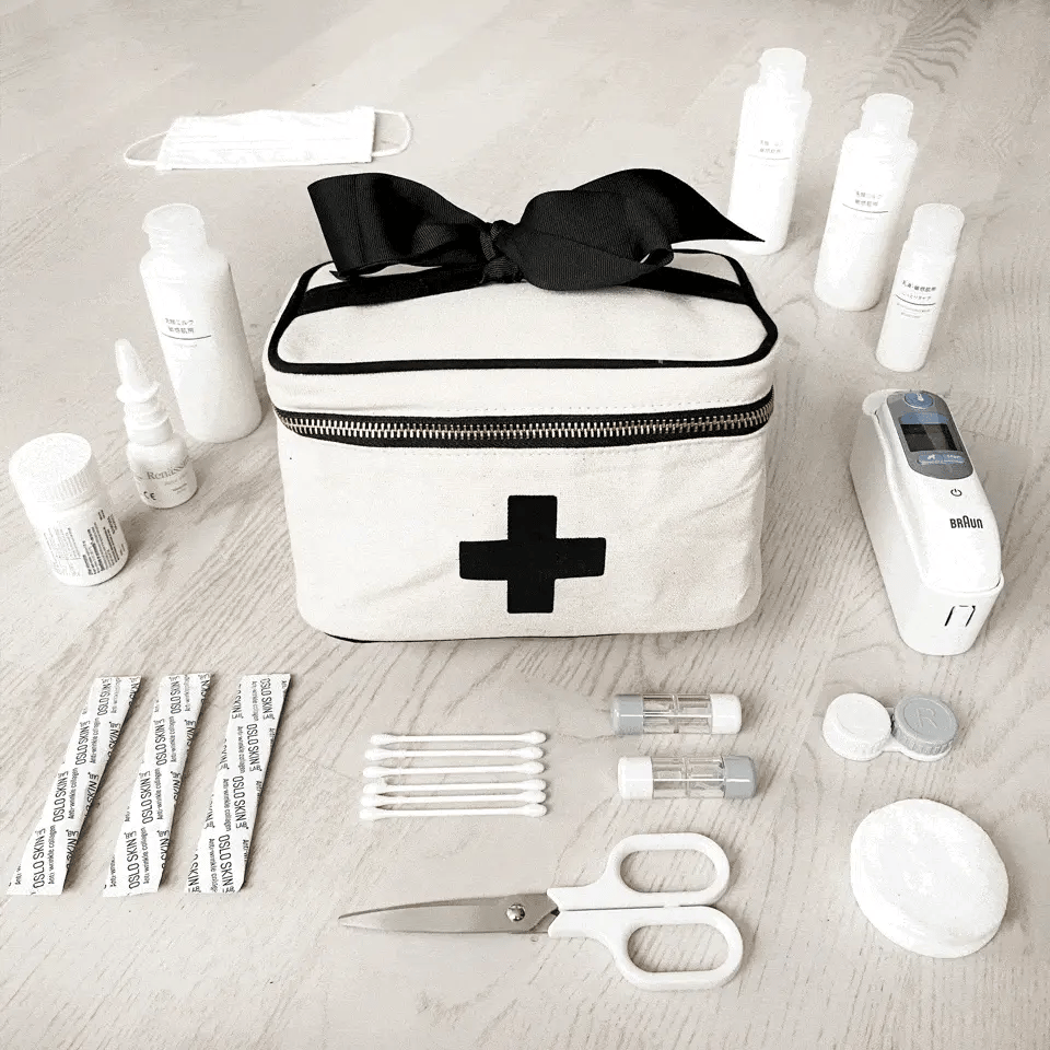 Bag-all Meds and First Aid Storage Box in cream with black cross design, black bow accent, and zipper closure, surrounded by medical supplies