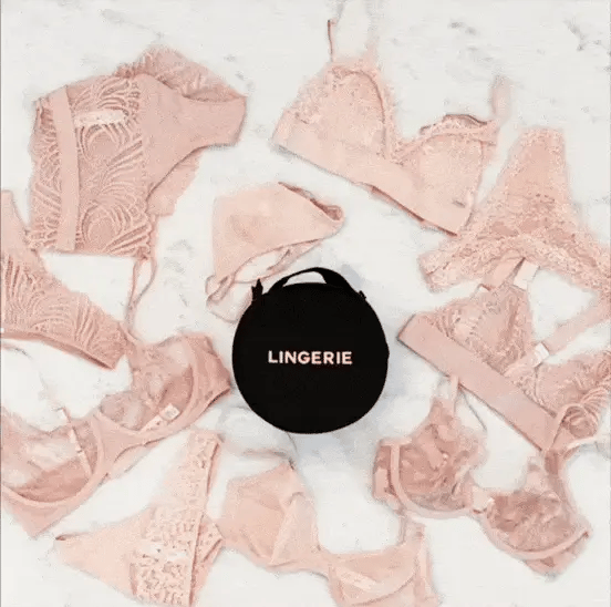 Bag-all Round Lingerie Case in Black surrounded by blush pink lace lingerie pieces arranged in a circular pattern on white marble surface, showcasing elegant storage solution