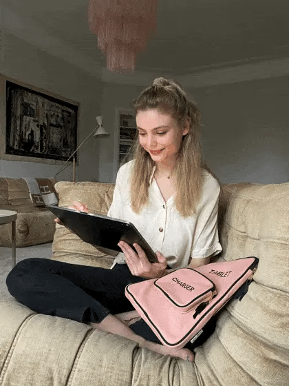 Bag-all pink tablet case with charger pocket shown on couch, featuring padded cotton design with front storage compartment and grosgrain ribbon detail in blush color