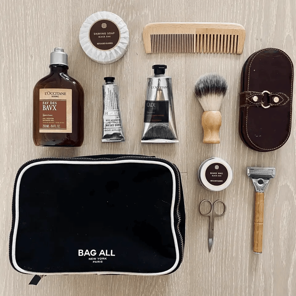Bag-all Double Sided Toiletry Case in Black shown with grooming essentials: shaving cream, wooden comb, after-shave balm, shaving brush, and manicure tools. Waterproof cotton case with dual zippers and white trim.