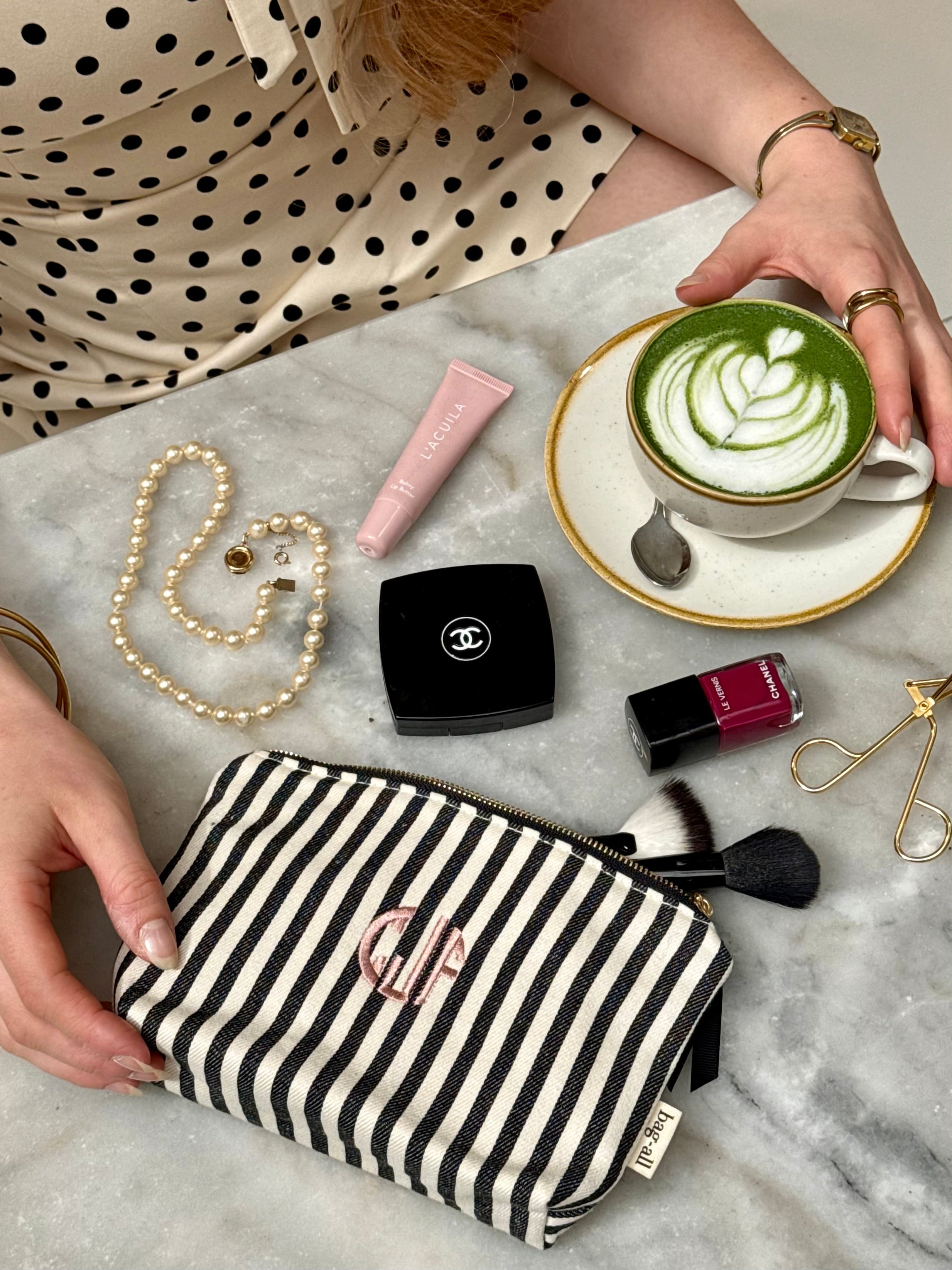 Striped cotton pouch with pink monogram, surrounded by makeup, pearl necklace, and matcha latte, showcasing Bag-all’s personalized travel organization style.