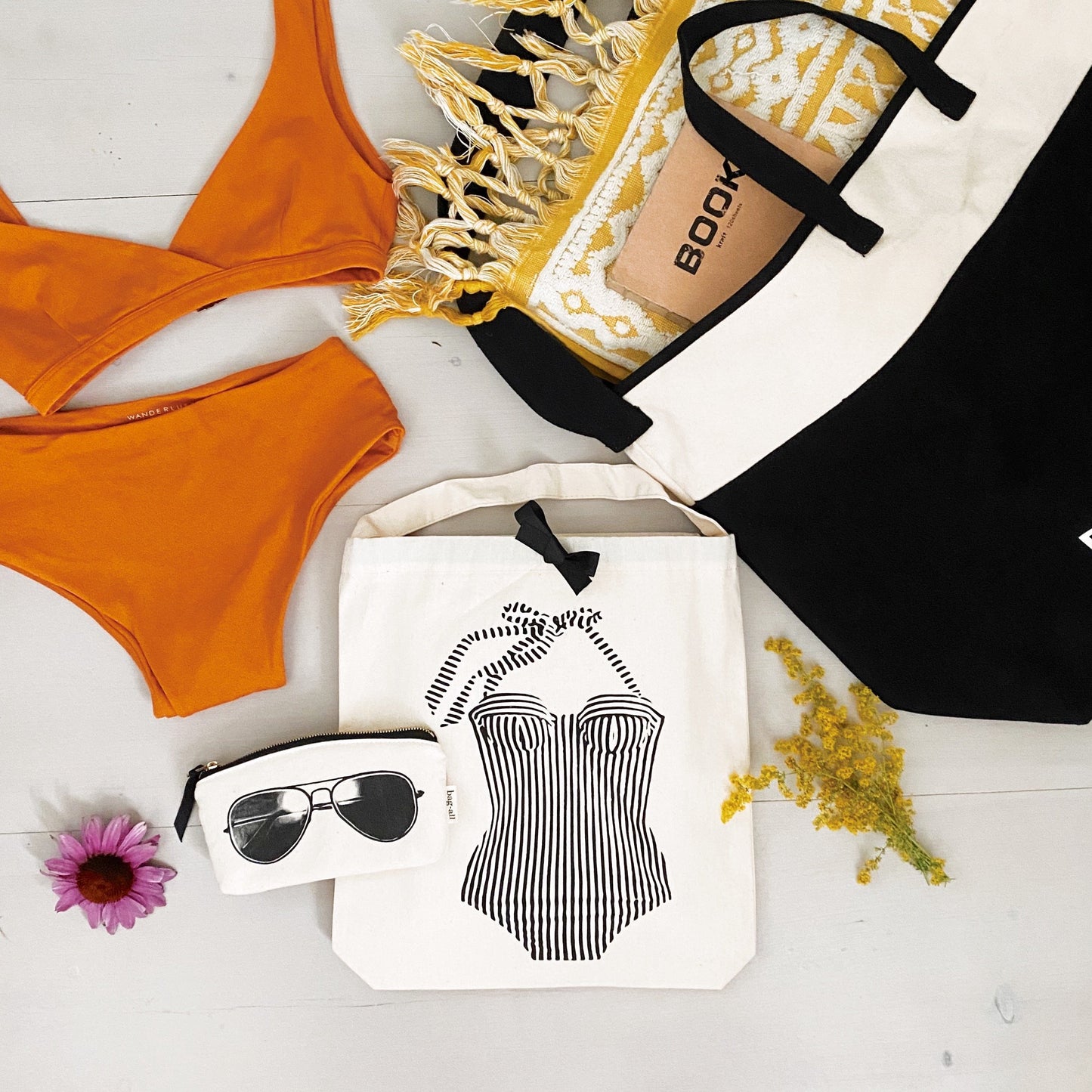 Bag-all Swimsuit Organizer Set with orange bikini, white cotton tote featuring striped swimsuit print, beach towel, sunglasses, and yellow flowers on white surface