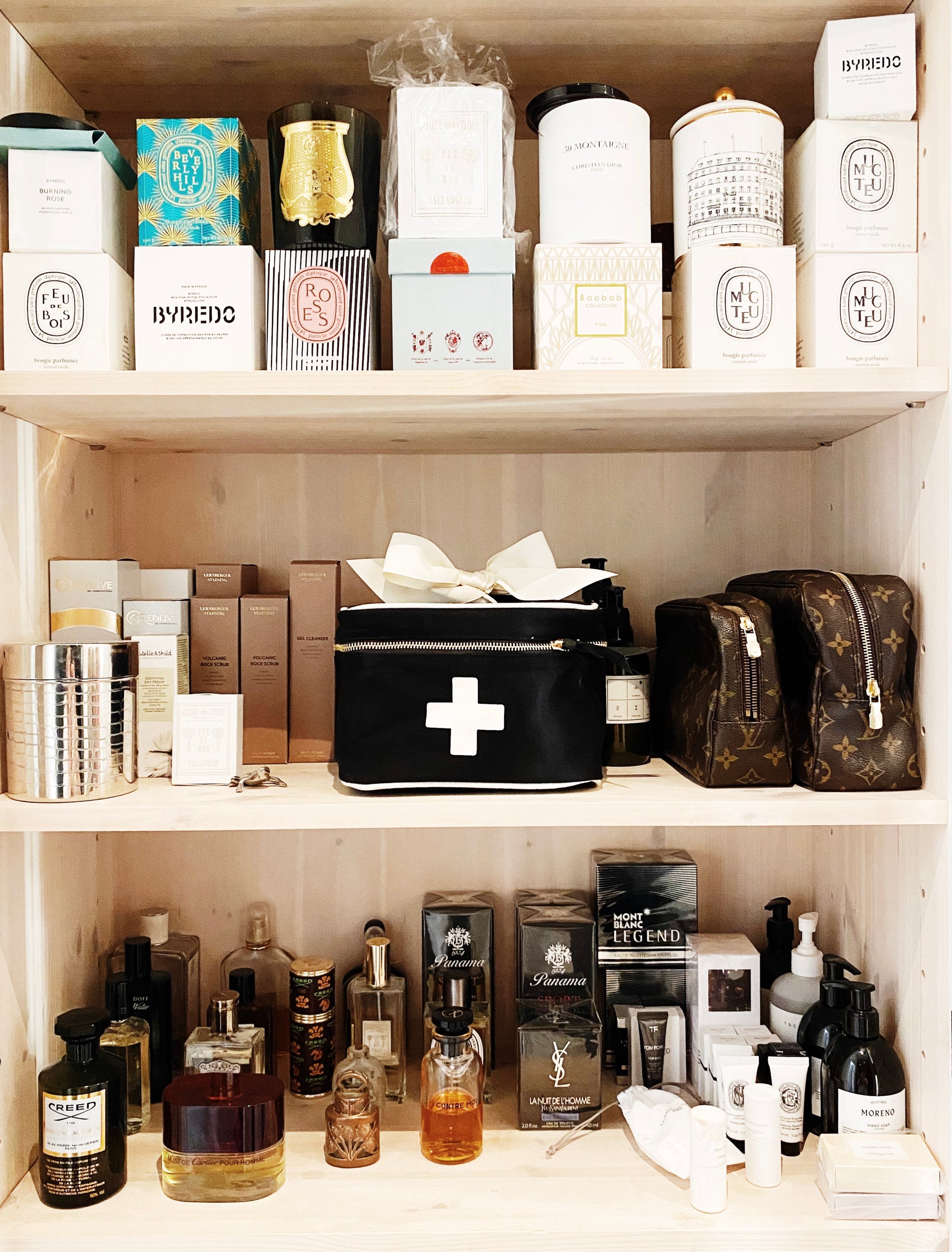 Bag-all Meds and First Aid Storage Box in black with white cross design, positioned on wooden shelf among luxury products and perfumes, features dual zippers and carrying handle