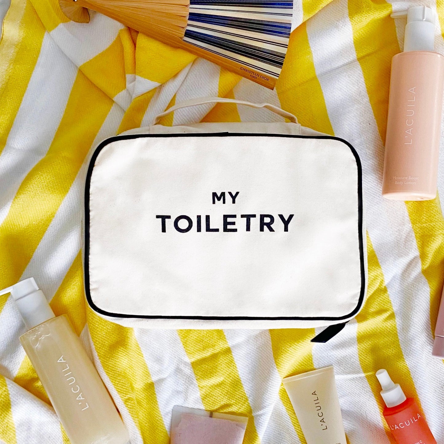 Bag-all Folding/Hanging Toiletry Case in cream with 'MY TOILETRY' text on striped yellow background, displayed with beauty products and makeup brushes