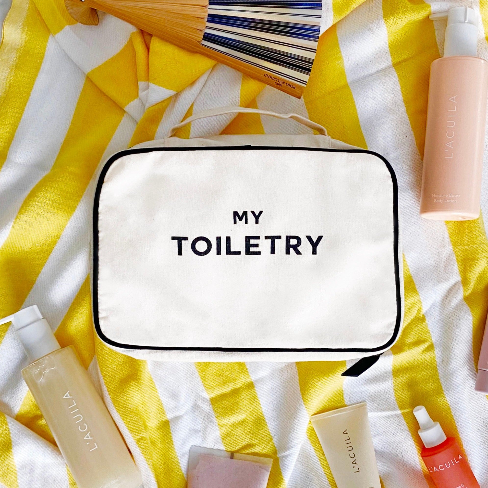 Bag-all Folding/Hanging Toiletry Case in cream with 'MY TOILETRY' text on striped yellow background, displayed with beauty products and makeup brushes