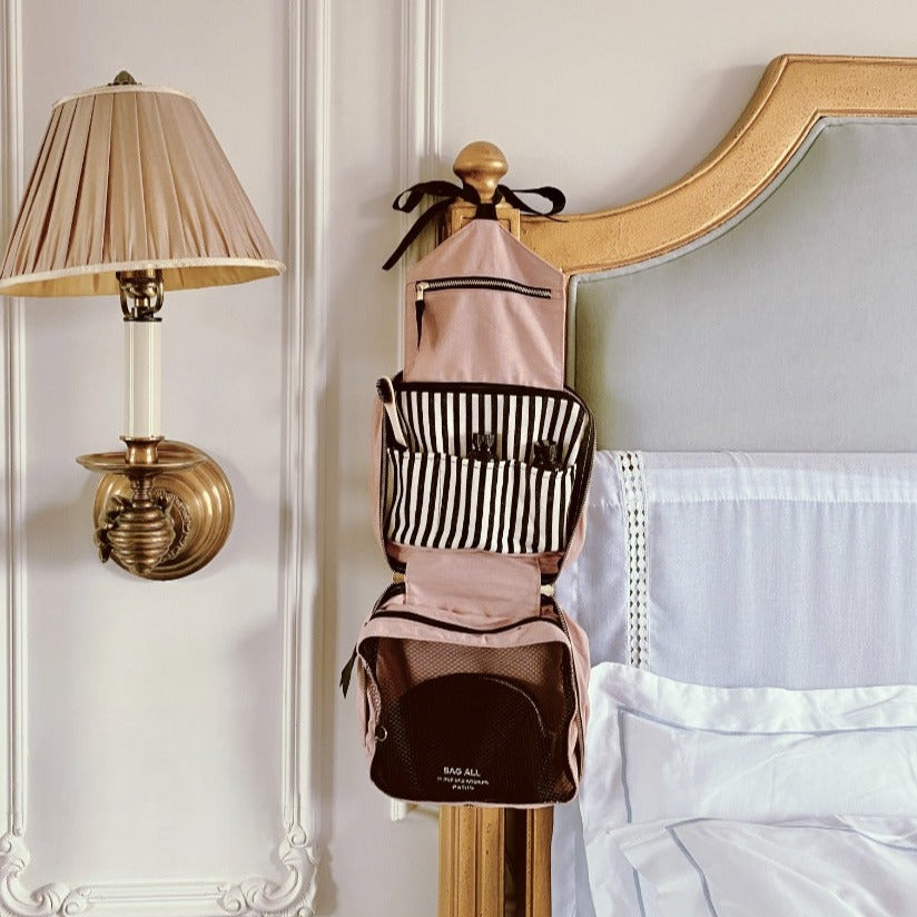 Bag-all Folding/Hanging Toiletry Case in blush pink with multiple compartments, striped interior, and hanging hook, displayed on elegant wall next to vintage-style lamp in bedroom setting