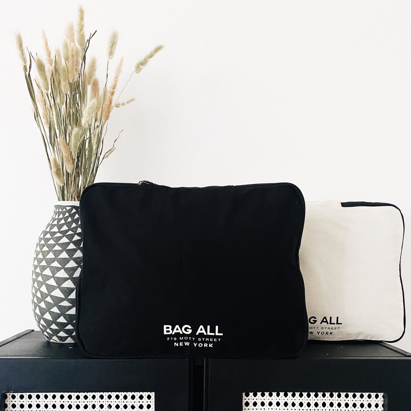 Bag-all Large Double Sided Packing Cube in Black, featuring dual compartments and brand logo, displayed next to geometric vase with dried pampas grass on black surface