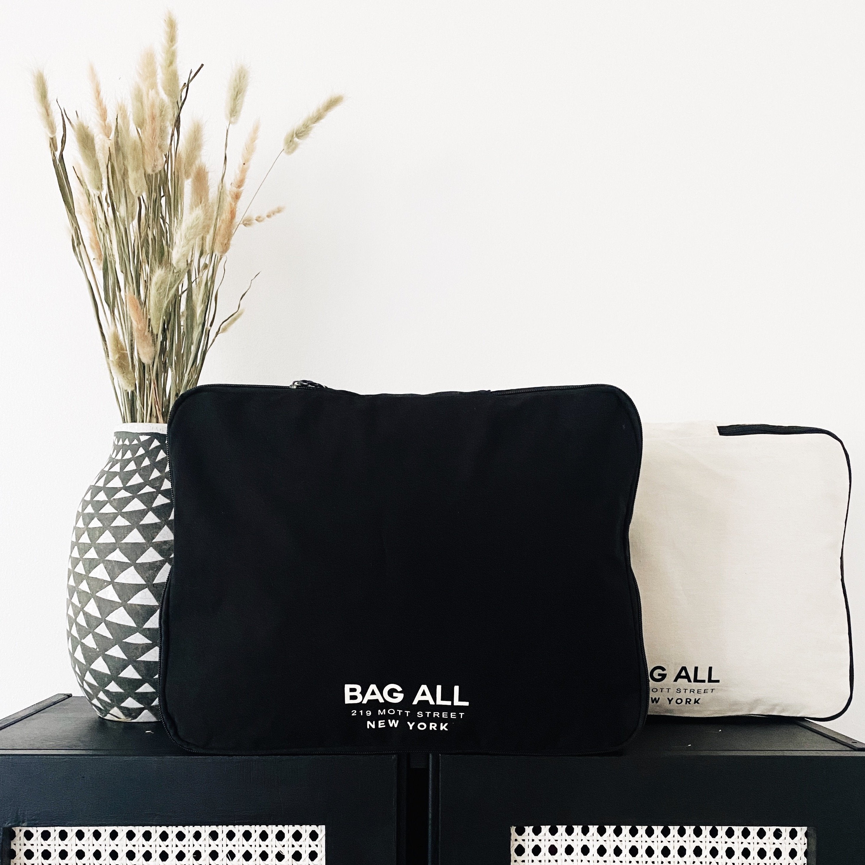 Bag-all Large Double Sided Packing Cube in Black, featuring dual compartments and brand logo, displayed next to geometric vase with dried pampas grass on black surface