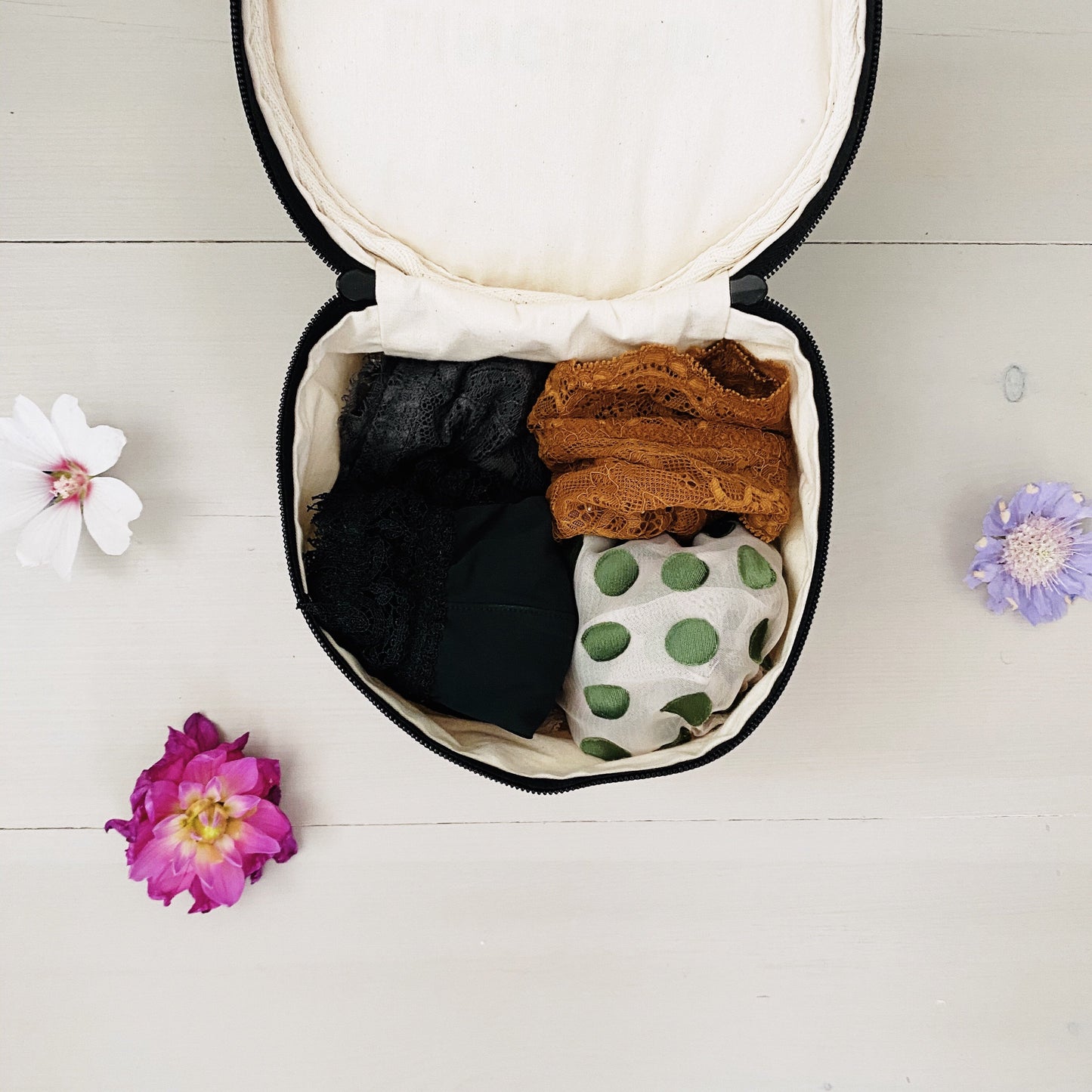 Bag-all Round Lingerie Case in cream color, opened to display neatly organized undergarments in black, brown, and polka dot patterns, surrounded by delicate flower petals on white surface