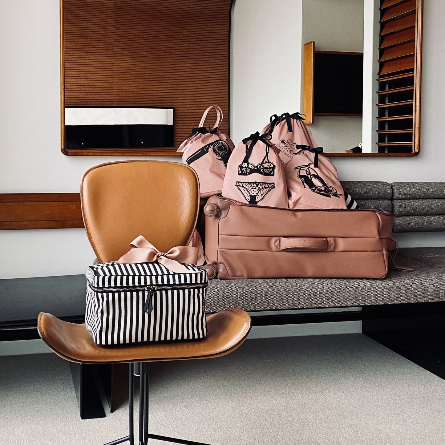 Bag-all Medium Box Makeup & Toiletry Striped case displayed on brown leather chair, featuring black and white stripes pattern, shown alongside pink travel bags in modern interior setting