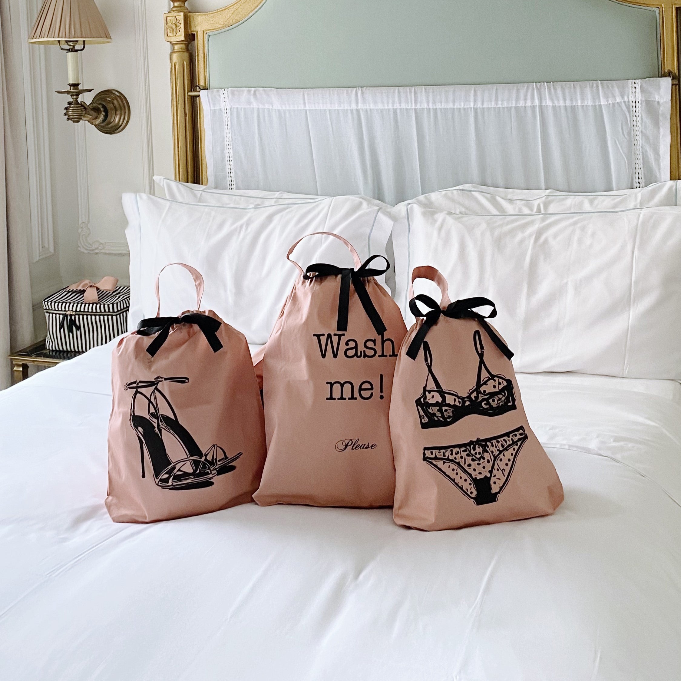 Bag-all pink polkadot lingerie travel bags featuring stylish black illustrations of shoes and undergarments with elegant black bow ties and text, displayed on white bedding against vintage headboard