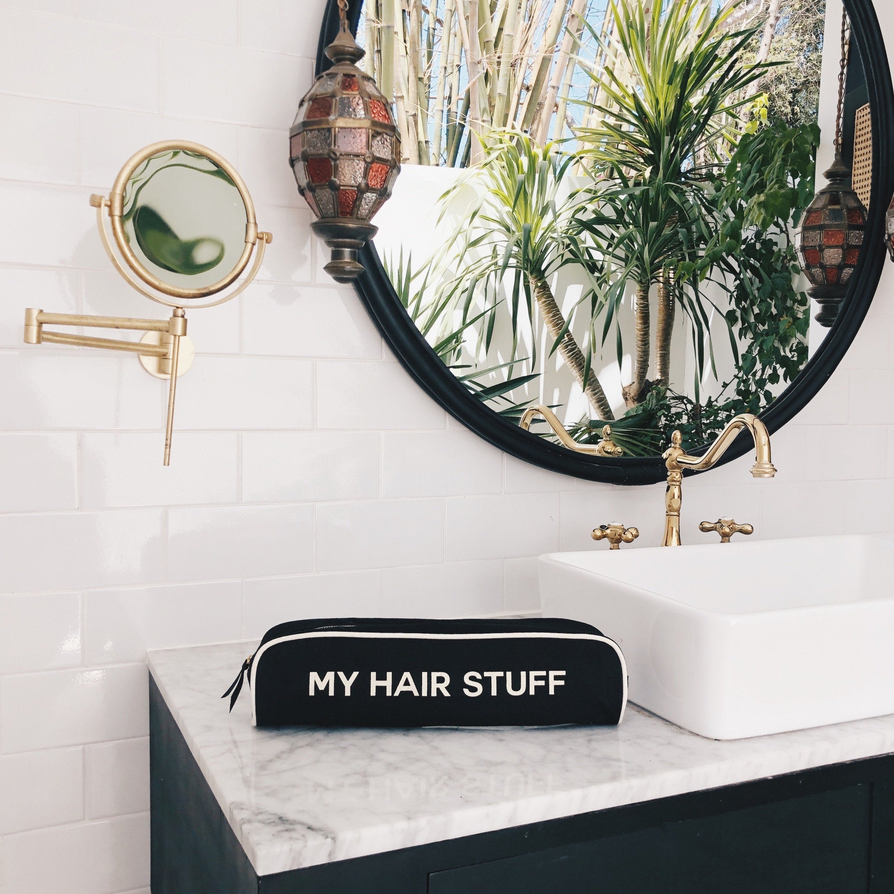 Bag-all Hair Stuff Travel Case in black displayed on marble bathroom counter, featuring elegant gold-trim mirror and tropical plants in background