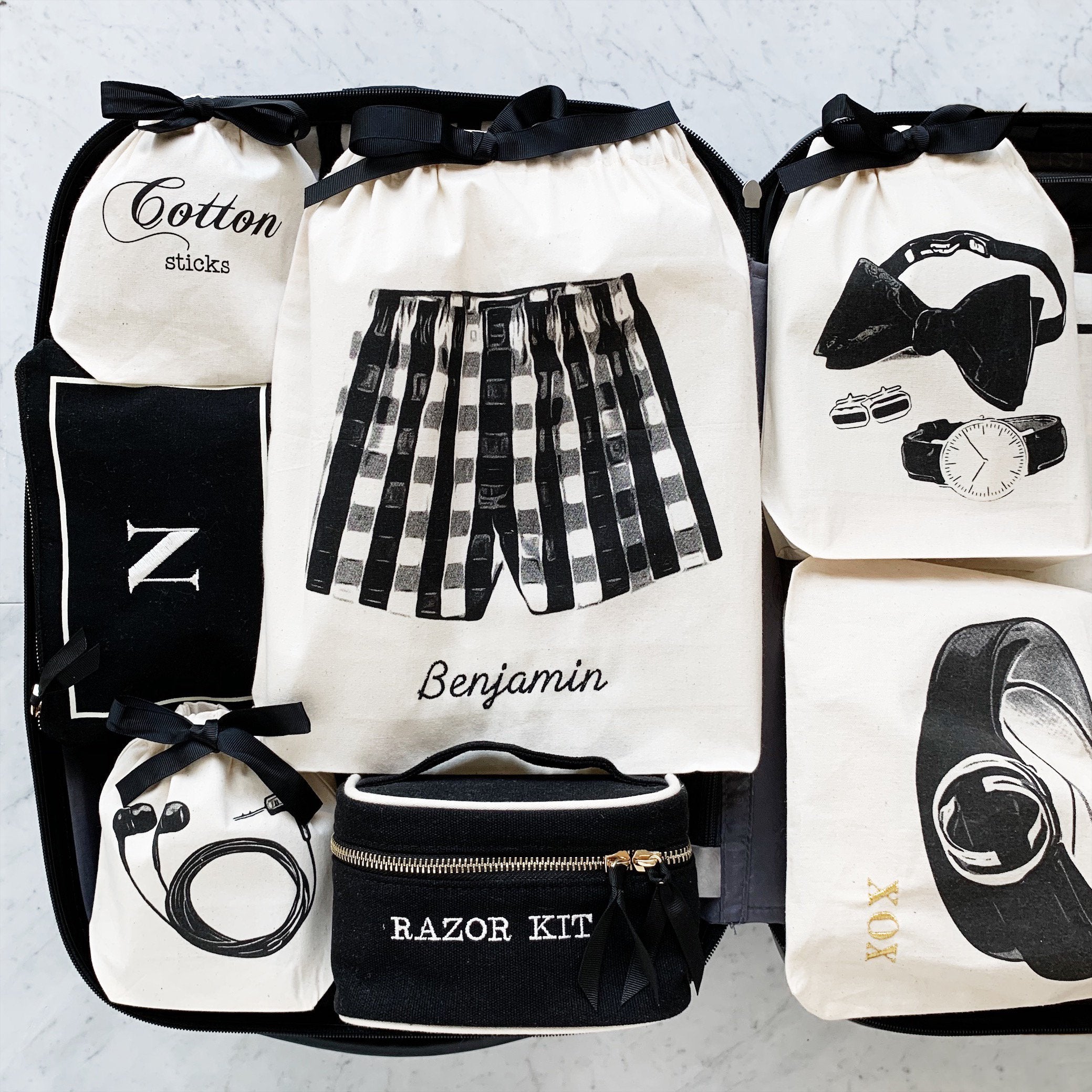 Bag-all travel organizer bags featuring monochrome illustrations of boxer shorts, bow tie, watch, and toiletries on cotton drawstring pouches with black ribbon accents