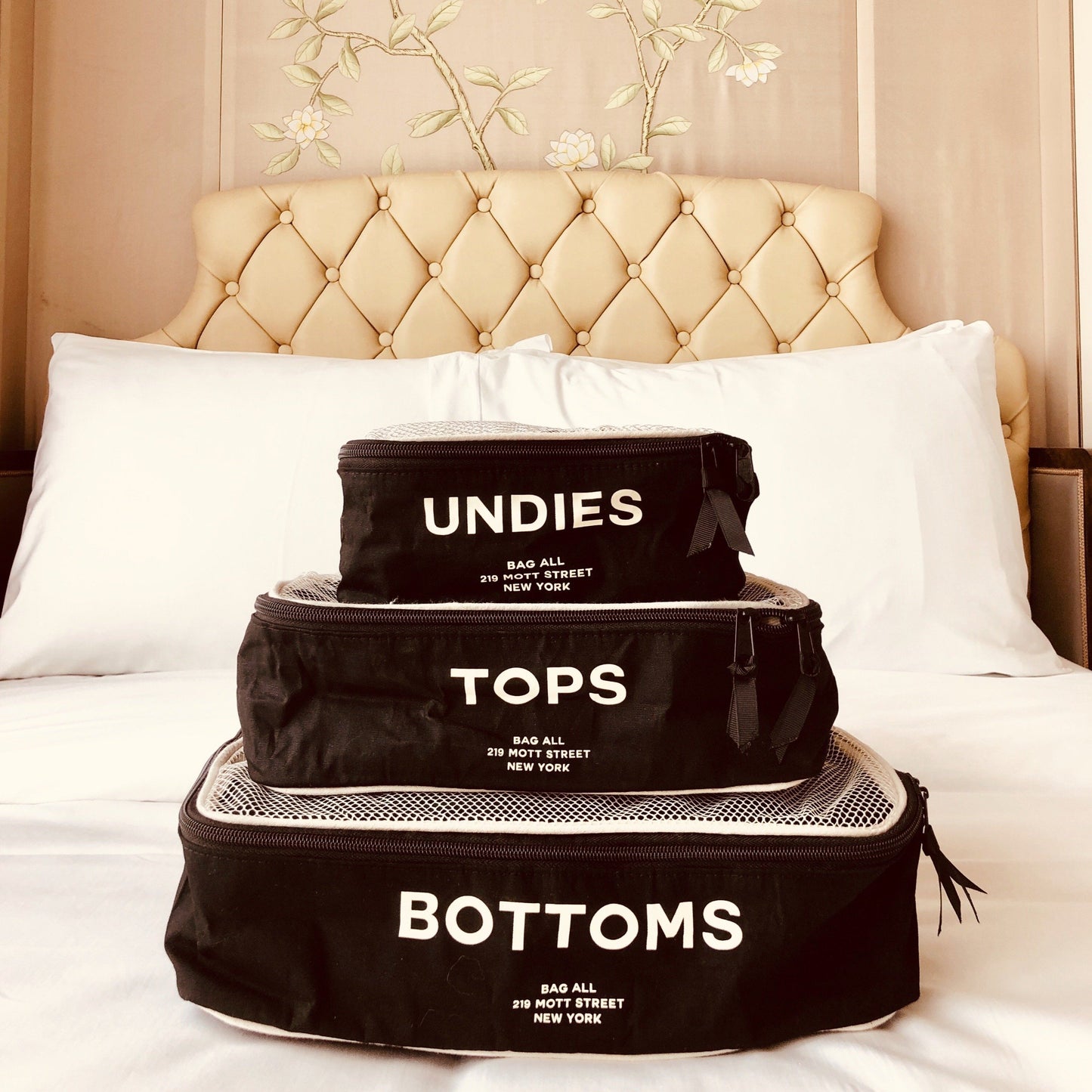 Bag-all Cotton Packing Cubes in black with white mesh top, stacked in three sizes labeled Undies, Tops, and Bottoms, displayed on white bedding against tufted headboard