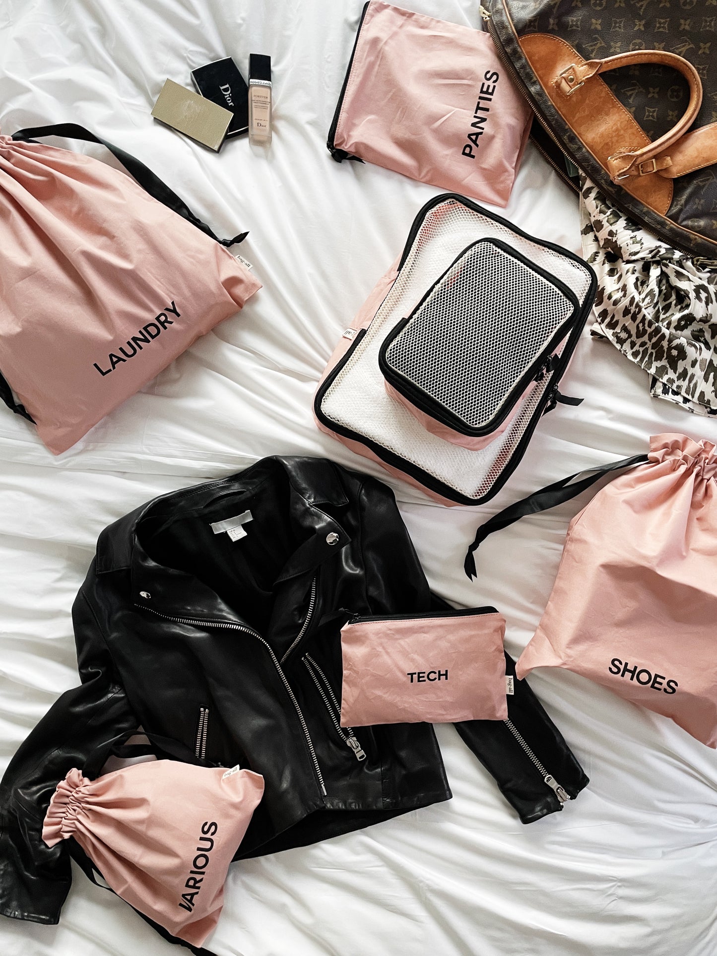 Bag-all travel organizer set featuring blush pink storage bags for laundry, tech, shoes, and panties, displayed with leather jacket and designer handbag on white bedding