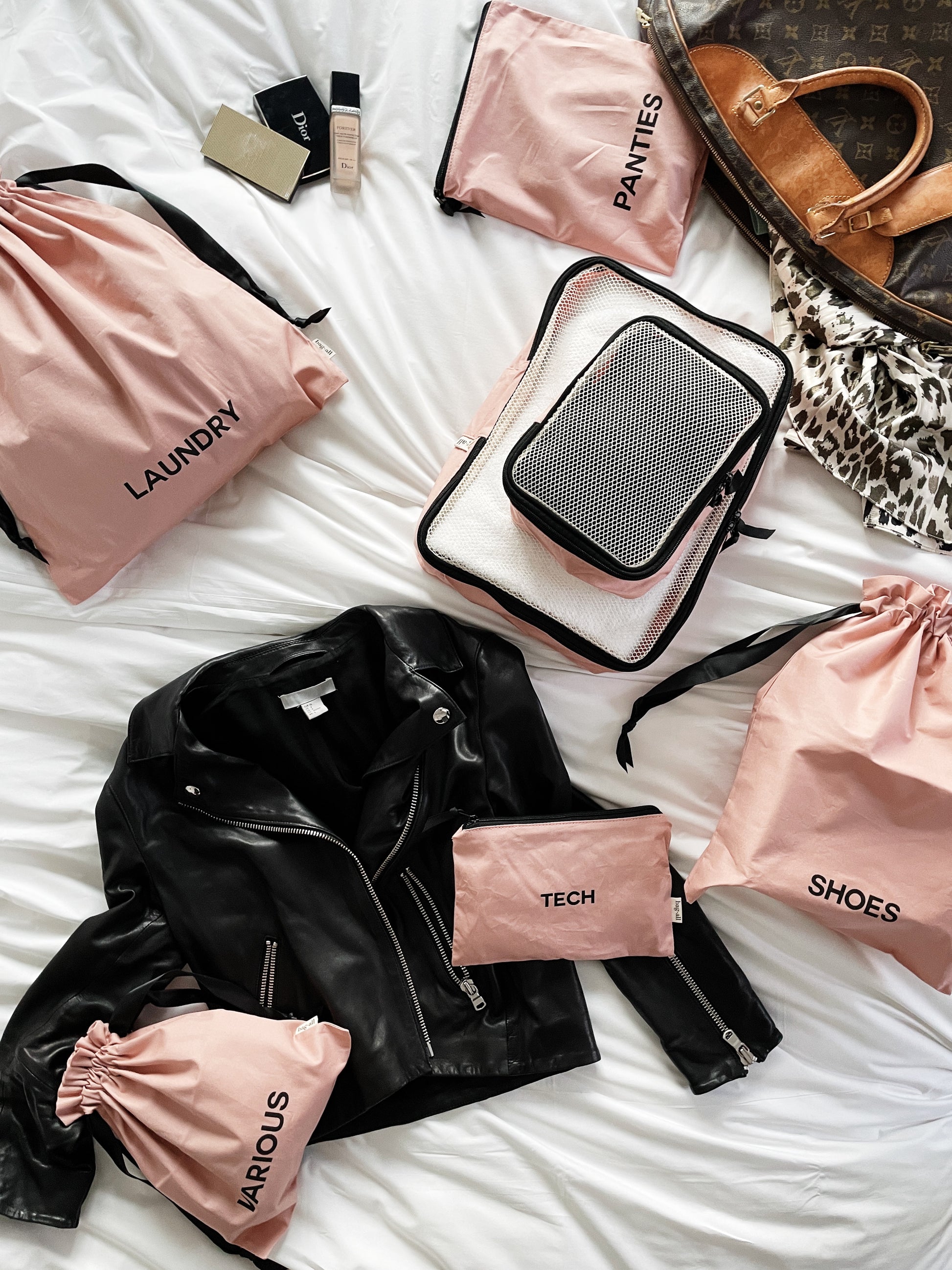 Bag-all travel organizer set featuring blush pink storage bags for laundry, tech, shoes, and panties, displayed with leather jacket and designer handbag on white bedding