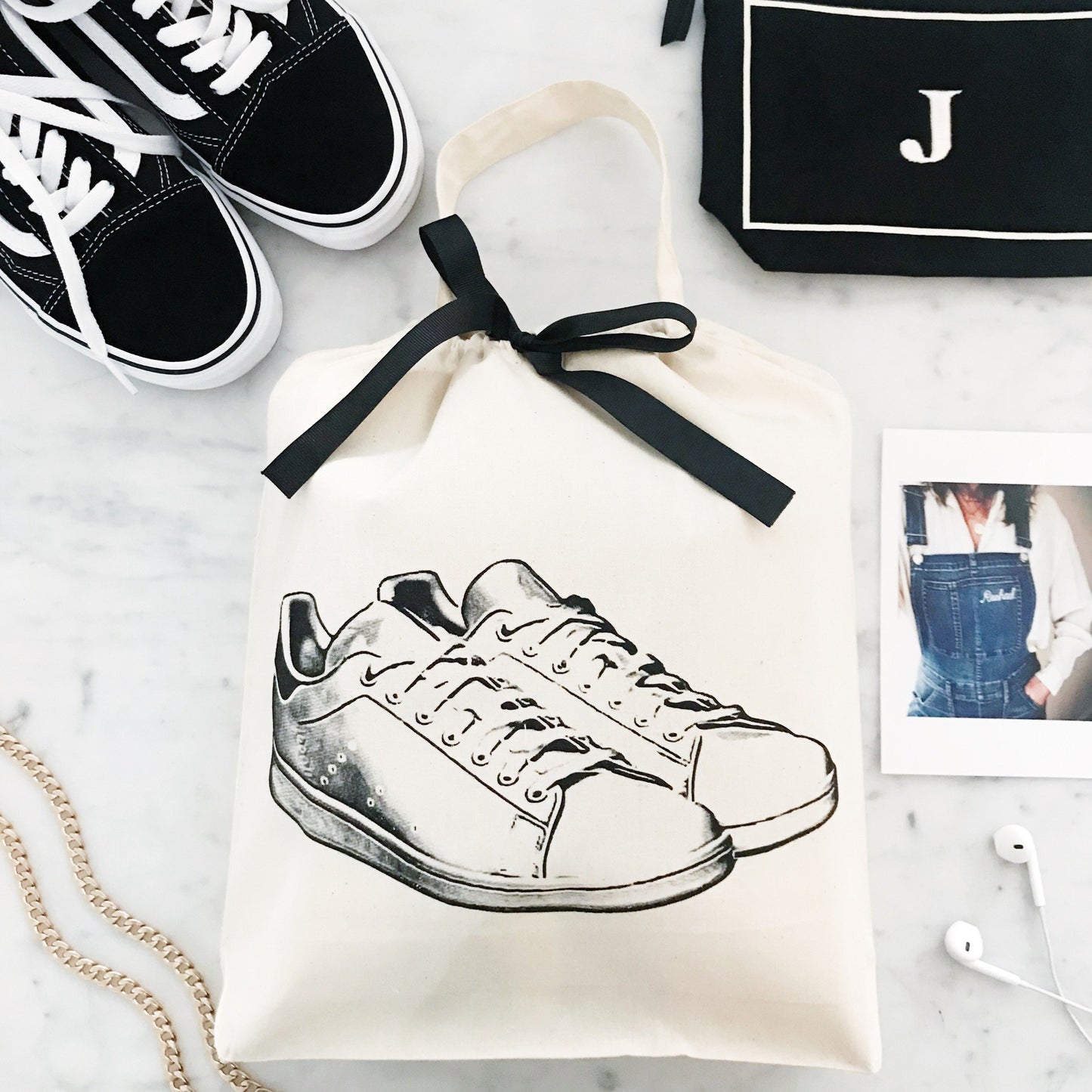 Bag-all White Sneaker Shoe Bag featuring hand-drawn sneaker design on cream cotton canvas with black ribbon tie, displayed alongside black sneakers and accessories on marble surface