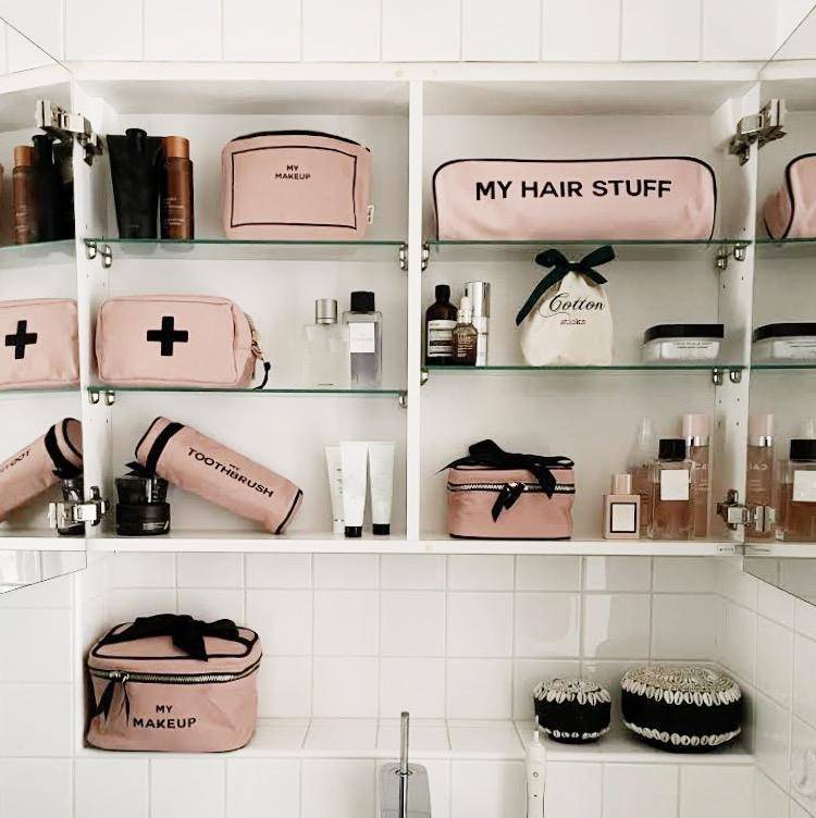 Bag-all My Makeup Pouch in blush pink with black text, styled on white bathroom shelves alongside matching pink organizers and beauty products, featuring gold zipper and coated lining